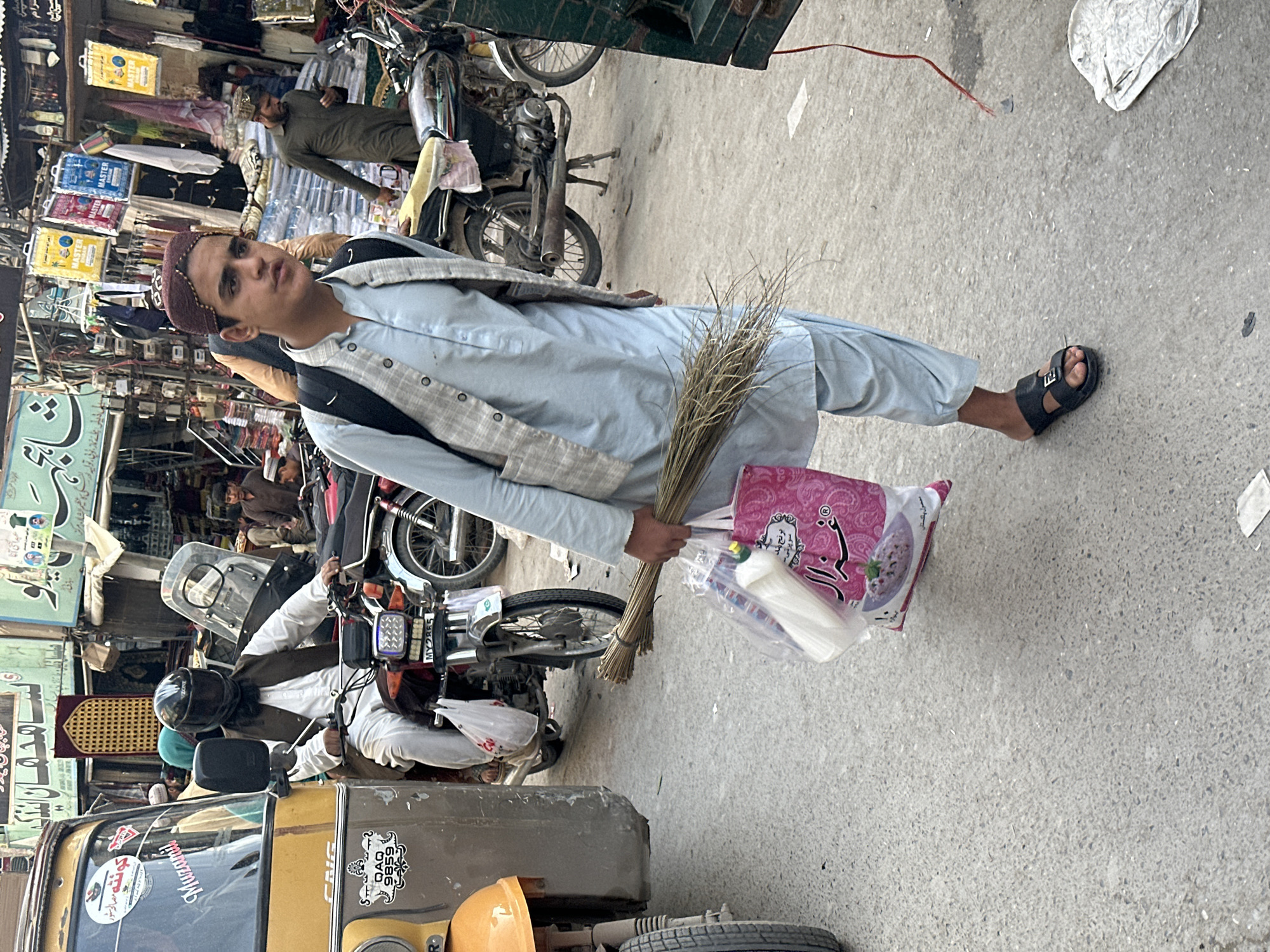 The image captures a vibrant, bustling street scene, likely a market in a South Asian country, during daylight hours.
The central figure is a young man, standing on a paved street and looking directly at the viewer. He is dressed in a light blue shalwar kameez, a grey and white patterned waistcoat, and a maroon embroidered prayer cap. He wears black open-toed sandals and has a black backpack. In his left hand, he holds a bundle of thin dried sticks (possibly for cleaning or traditional toothbrushes) and a pink shopping bag containing a clear plastic package with white items, likely tissues or napkins, and another small object.
The background is rich with activity and details. Several motorcycles are visible; one man, wearing a dark green outfit and a cap, is seated on a motorcycle directly behind the main subject. Further left, another individual in a white outfit and a black helmet is seen on a motorcycle, which appears to be attached to a three-wheeled vehicle or part of an auto-rickshaw, bearing the license plate "MY2865". A yellow and brown auto-rickshaw is partially visible in the bottom-left, with "Muzamil" and "QAQ 9859" written on its side.
The street is lined with market stalls and shops, featuring various items for sale. Yellow and blue packages labeled "MASTER" are prominently displayed. Walls are adorned with posters and signs, some featuring Urdu or Arabic script. The ground is a light-colored concrete or asphalt, with scattered debris including a white plastic bag and a red string.
Visible text includes "MASTER" on product packaging, "MY2865" on a motorcycle license plate, "Muzamil" and "QAQ 9859" on the auto-rickshaw. Other signs have Urdu/Arabic script, such as "کونته مصالح سشور" and parts of another sign reading "ی کا عظیم الشان مرکز نماز، نوبی تولید". The pink shopping bag also displays "برج" (Burj) and a product image. The overall impression is that of a typical, lively market day.