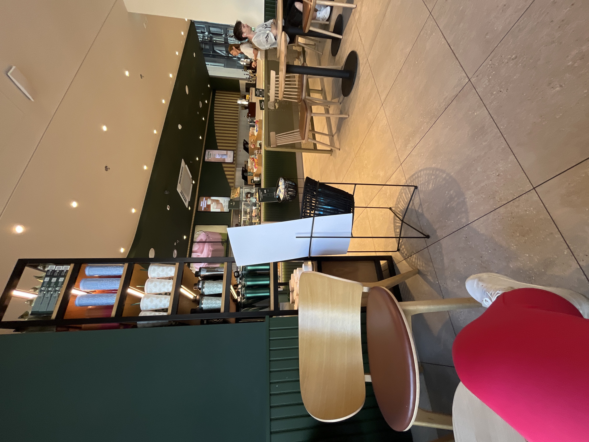The image depicts the interior of a cafe. In the foreground, a person's legs, clad in bright red leggings and white sneakers, are visible as they sit in a chair with a brown seat and wooden frame. To the left, a dark green wall with vertical wooden paneling is present, alongside a tall shelving unit displaying various blue, white, and green cylindrical cups, as well as black canisters. In the mid-ground, another wooden chair with a brown seat is positioned near a black metal stand holding a white board and a black basket. Further back, several tables with wooden chairs are set up. In the background, a counter area is visible with baristas and customers, and a large window reveals an outdoor scene. The flooring throughout the cafe appears to be large, light-colored tiles. The overall lighting is bright, suggesting daytime.
