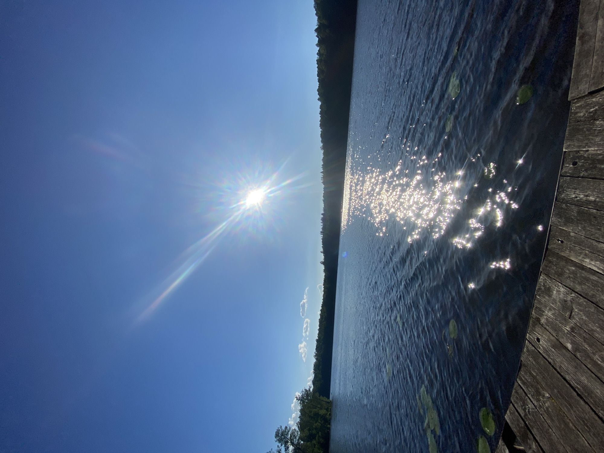 A bright sun shines in a clear blue sky over a calm lake, casting shimmering reflections on the water's surface. The sun's rays create a starburst effect with a rainbow halo. A dense line of dark green trees forms the horizon along the far side of the lake, with a few scattered white clouds in the sky. In the foreground, a wooden dock or pier extends into the water, showing weathered planks with visible grain and nail holes. Lily pads float on the dark blue water near the dock. The overall scene depicts a serene natural environment. The location is described as the Азёрскі сельскі Савет in Belarus. There are no people or animals visible in the image.
