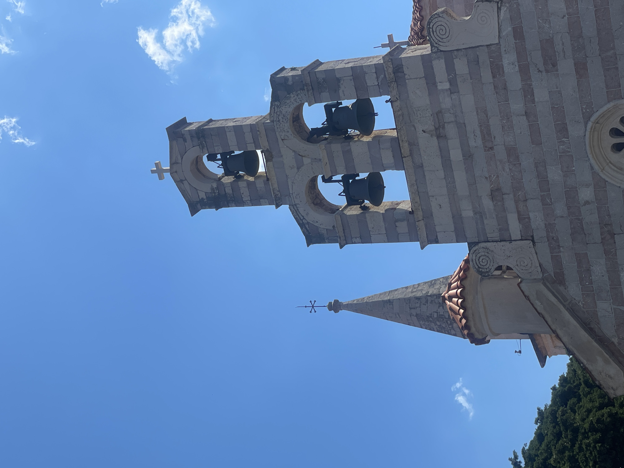 The image captures a low-angle view of a stone church tower, likely in Unknown, Unknown. The tower features a multi-tiered structure with arched openings housing bells. A cross adorns the tower's peak. The stonework exhibits alternating light and dark bands. Adjacent to the main tower, a smaller spire rises, topped with a decorative finial. Red tile roofing is visible on a connecting structure. A clear blue sky serves as the backdrop, with a few scattered clouds. Green foliage peeks into the frame from the lower right corner. The image is oriented vertically, with the church tower angled diagonally.