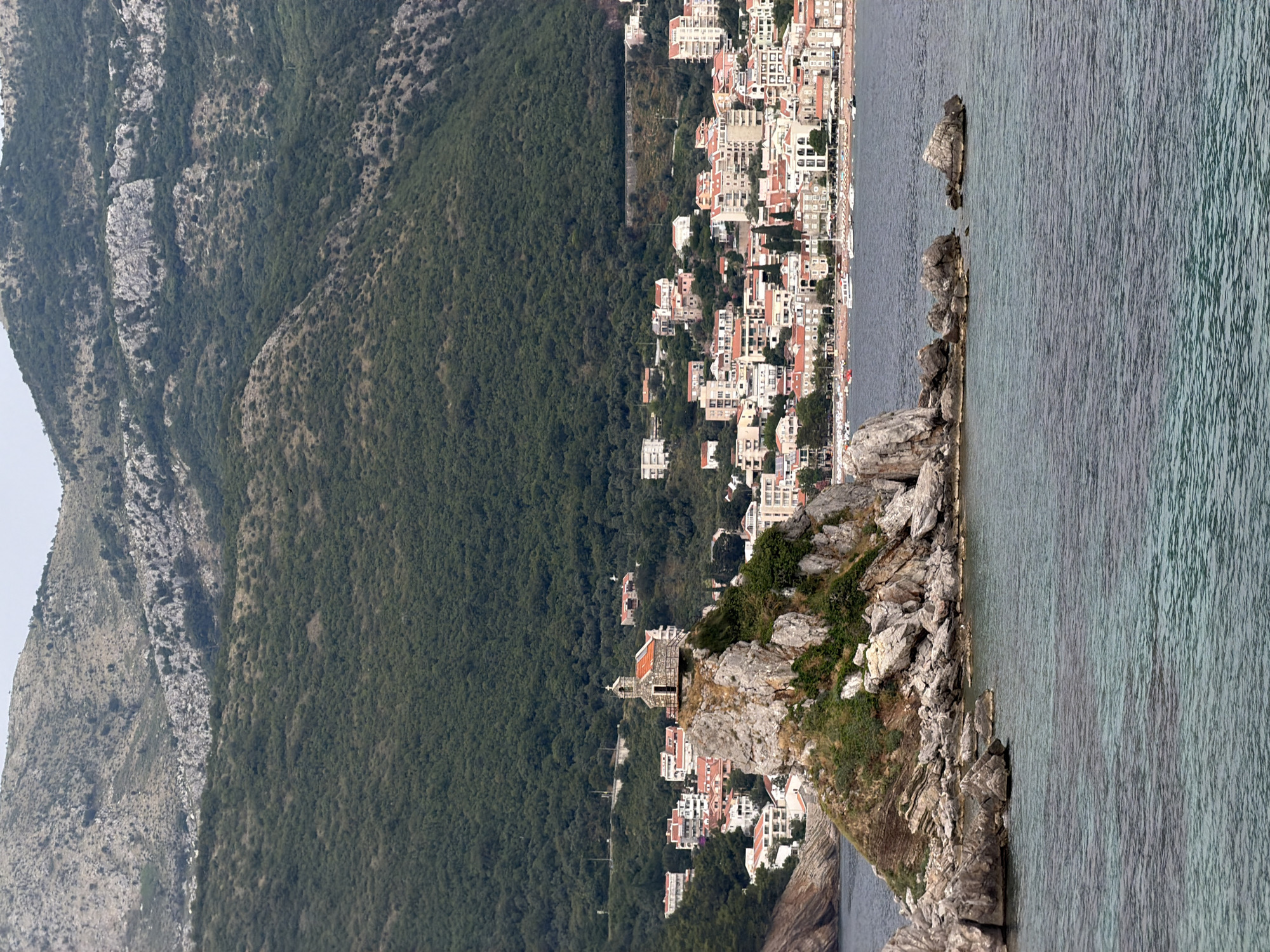 This media file depicts a scenic view of Petrovac na Moru, Montenegro. The scene is a coastal landscape featuring a densely forested mountainside on the left, descending towards a town nestled along the coastline. The town is characterized by numerous buildings with red-tiled roofs, tightly packed together, suggesting a vibrant urban area.
In the foreground, on the right side of the image, is the sea. The water appears calm, with a textured surface indicating gentle ripples. Several large rocks jut out from the water and along the coastline, adding to the rugged beauty of the location. A small, stone structure, possibly a church or chapel, with a red roof, is perched atop a rocky outcrop overlooking the sea and the town.
The overall impression is of a sunny day, likely late afternoon given the soft lighting. The lush greenery of the mountain and the clear blue of the sea create a harmonious and picturesque setting. The architecture of the town, with its dense arrangement of buildings and prominent church, provides clues to the local culture and history. No people are clearly visible, and no text is present in the image. The view evokes a sense of tranquility and natural beauty, characteristic of a Mediterranean coastal town.