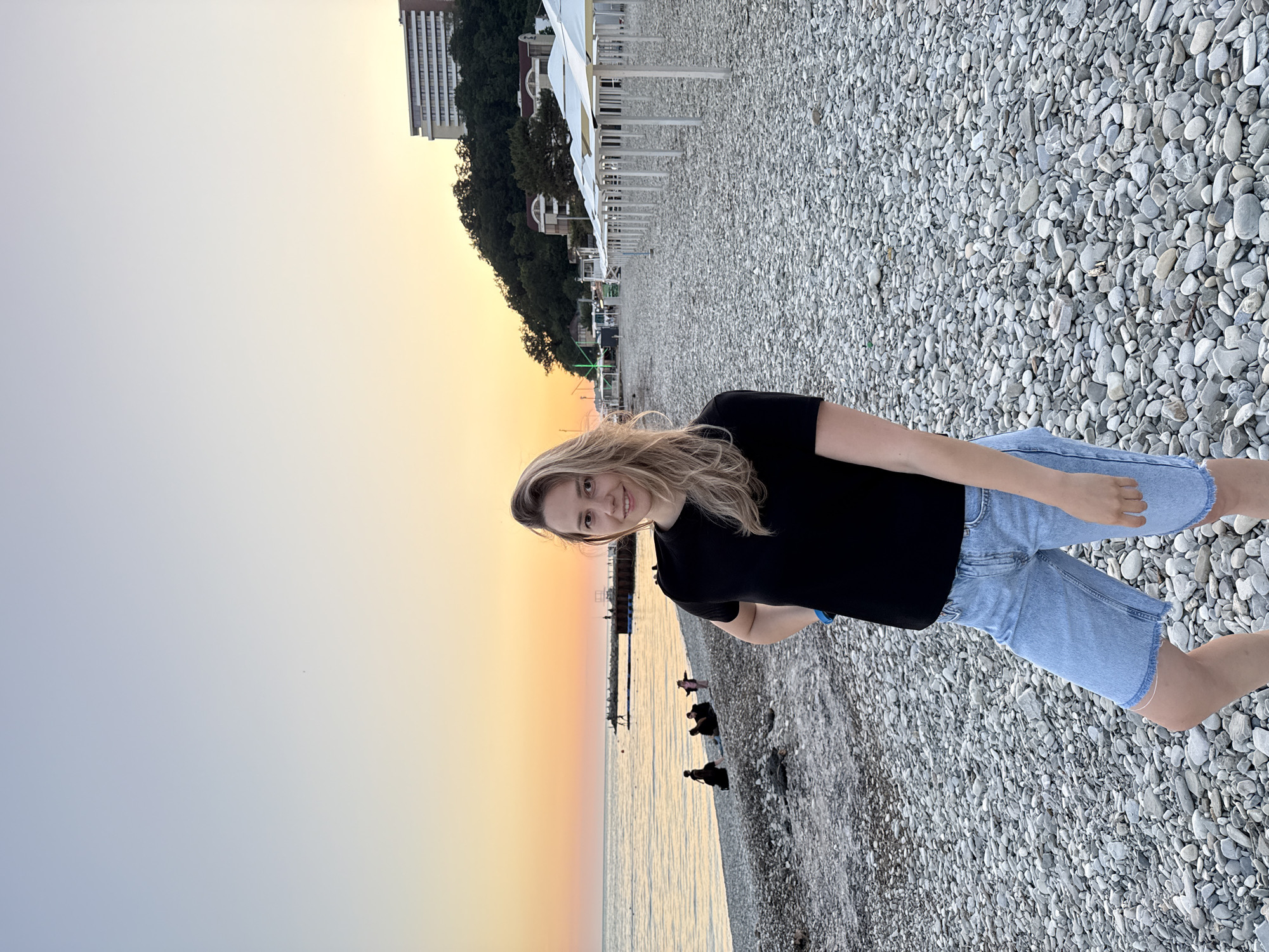 A medium shot photograph shows a woman standing on a pebble beach at dusk. The woman, with blonde hair and wearing a black t-shirt and denim shorts, is facing the camera with a slight smile. Her left hand is on her hip. In the background, the sea meets the sky in a gradient of orange and pale blue, indicative of a sunset. A pier extends into the water, with a few figures visible on it and along the shoreline. Further back, trees cover a hillside, topped by a tall, white hotel building. Rows of white beach umbrellas are set up on the pebble-covered beach. The scene is located in the city of Nebug, Russia.