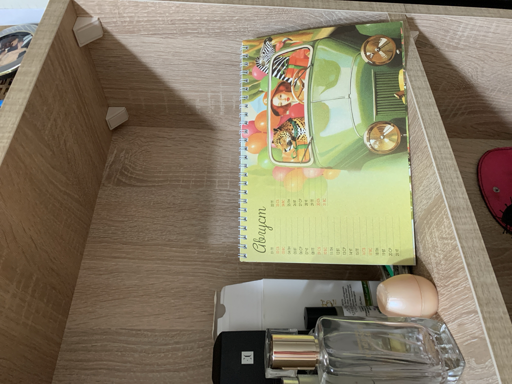 On a wooden shelf, a spiral-bound calendar featuring a cheerful illustration of a woman driving a green vintage car with a leopard and a zebra as passengers is displayed. The calendar's pages are open to the month of August, with dates and days of the week listed in Russian. To the left of the calendar, several perfume bottles are arranged, including a tall clear bottle with a metallic cap and a smaller, pale pink egg-shaped container. The background consists of the wood grain of the shelf and the vertical surface of a cabinet. The lighting suggests an indoor setting, possibly a home or retail environment. There is no visible information to determine its location in the city of Kapyĺ, Belarus.