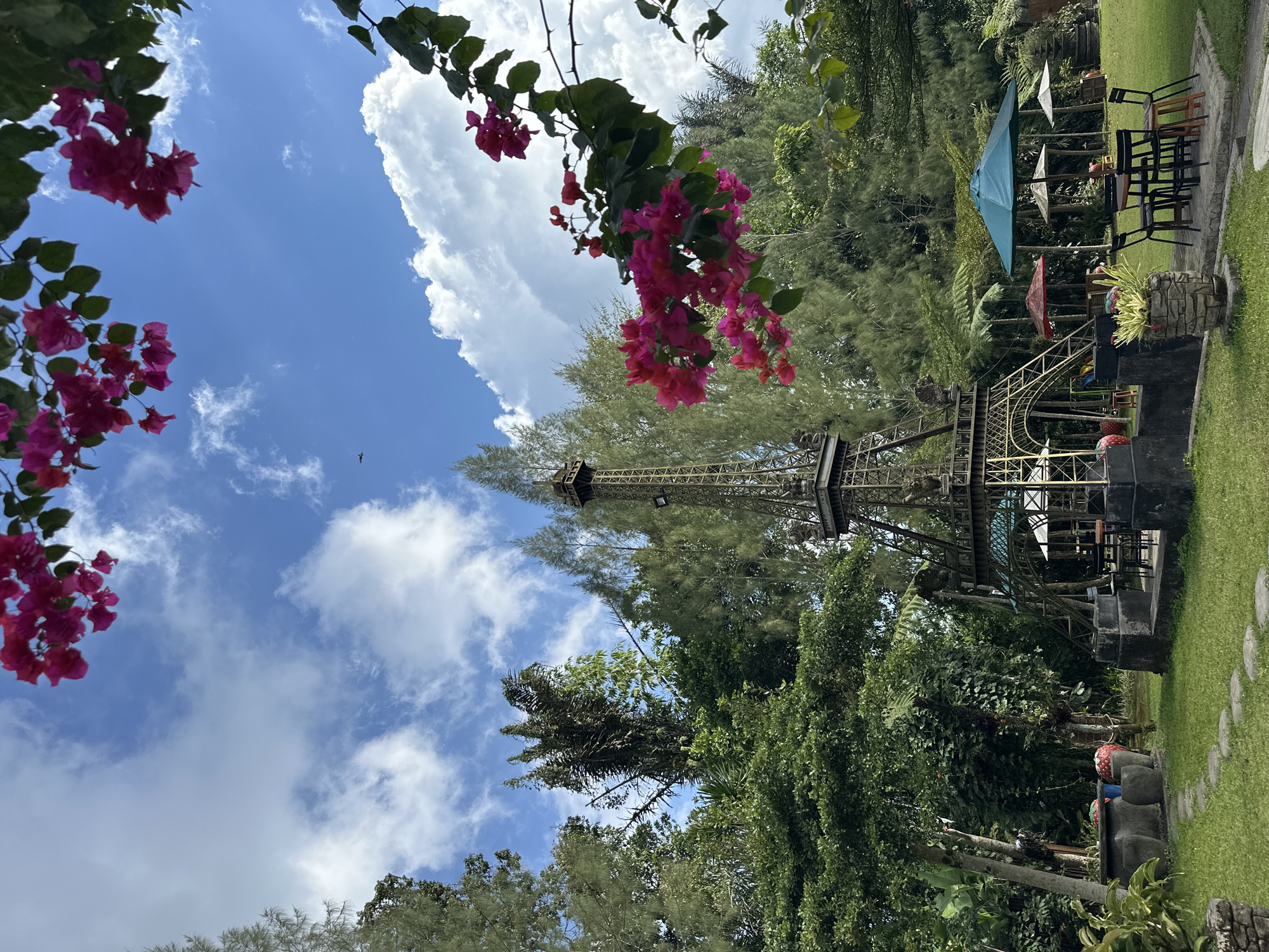 The image depicts an outdoor setting, likely a garden or park, with a miniature replica of the Eiffel Tower as a central focal point. The replica is made of dark metal and stands on a stone base. Surrounding the tower are various trees and lush green vegetation, including tall pines and smaller leafy plants. Several outdoor tables and chairs are arranged in front of the tower, some covered by umbrellas. A stone pathway made of stepping stones winds through the grassy area. In the foreground, there are large stone planters and decorative elements. The sky above is blue with scattered white clouds, and bright pink bougainvillea flowers are visible in the upper portions of the frame, framing the scene. In the distance, a small airplane is visible in the sky. The scene appears to be located in the city of Bangli, Indonesia, suggesting a themed garden or tourist attraction. There are no people visible in the image.