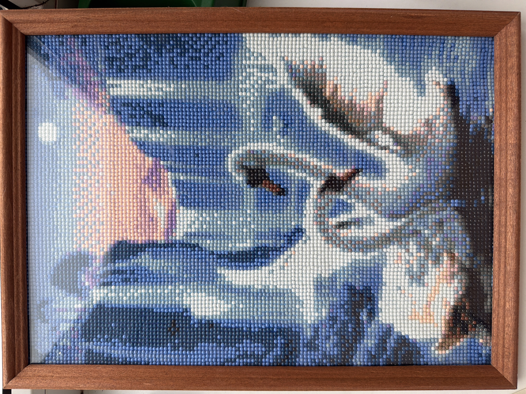 A framed diamond art picture depicting a scene with predominantly blue, white, and peach/pink hues. The artwork appears to show an abstract image, possibly of a landscape or cityscape with a light source on the left and darker elements on the right. The frame is wooden with a simple, clean design.