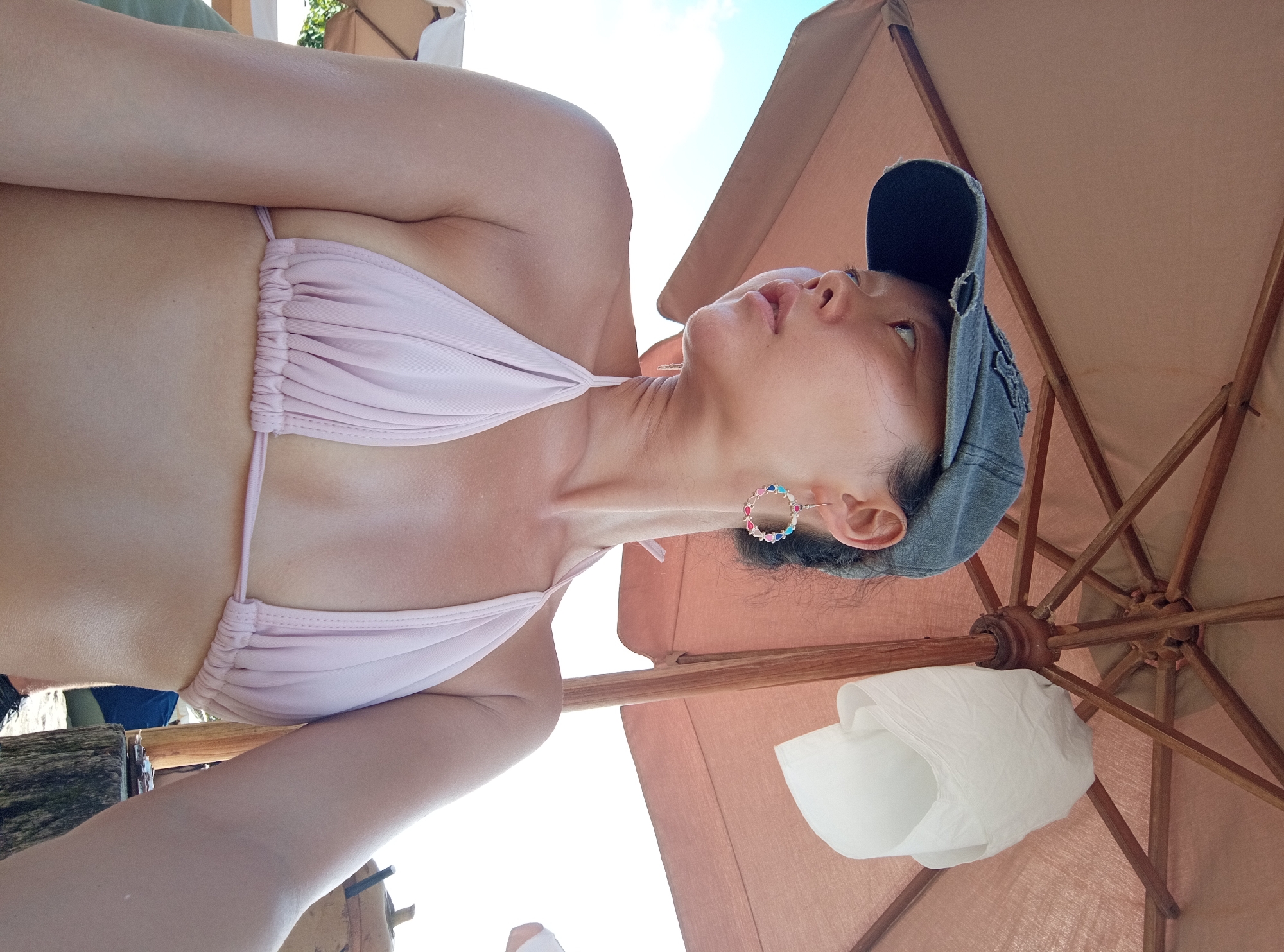 A woman wearing a light pink bikini and a dark-colored baseball cap is reclined under a large, brown patio umbrella. She is looking up and slightly to her left. She has a colorful, circular earring in her left ear. White fabric is tucked into the underside of the umbrella. Part of a wooden structure is visible at the lower left. The sky is visible in the background. The image was taken in North Kuta, Indonesia.
