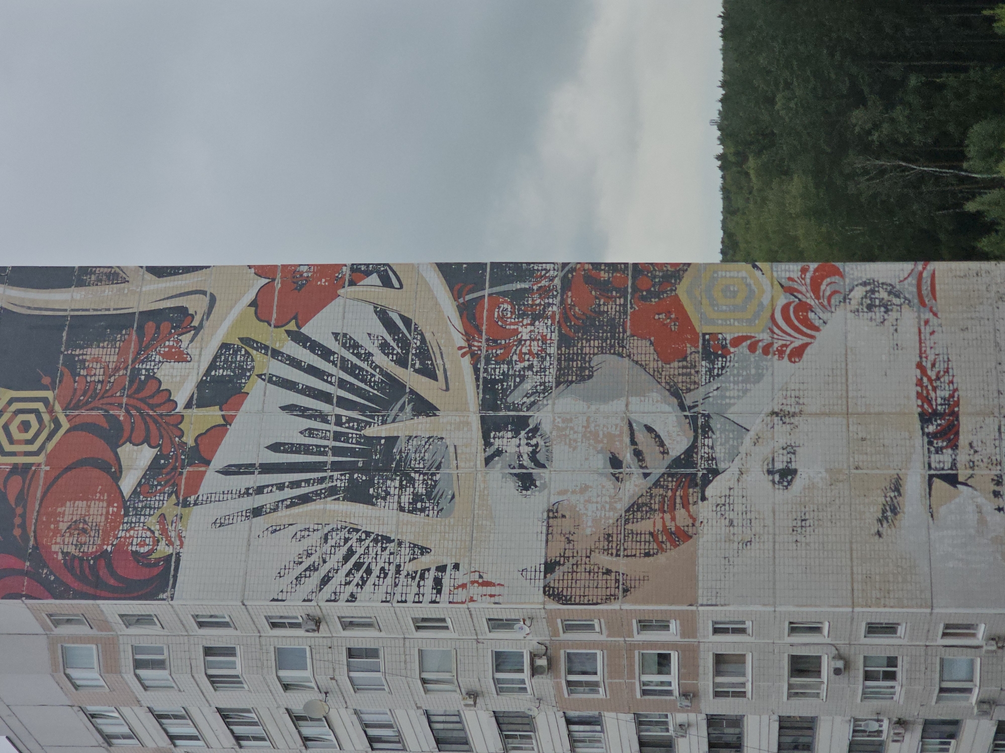 The image depicts a mural on the side of a residential building in Odintsovo, Russia. The mural features a stylized portrait of a woman with a crown or headdress made of spiky shapes and floral elements. The artwork is rendered in a graphic style with bold colors like red, black, and yellow, contrasted against a pale background. The building itself is a multi-story, light-colored apartment block with numerous windows and air conditioning units visible. Below the building, a dense forest stretches across the horizon under a cloudy, overcast sky. The overall scene is a juxtaposition of urban architecture and large-scale street art against a natural backdrop.