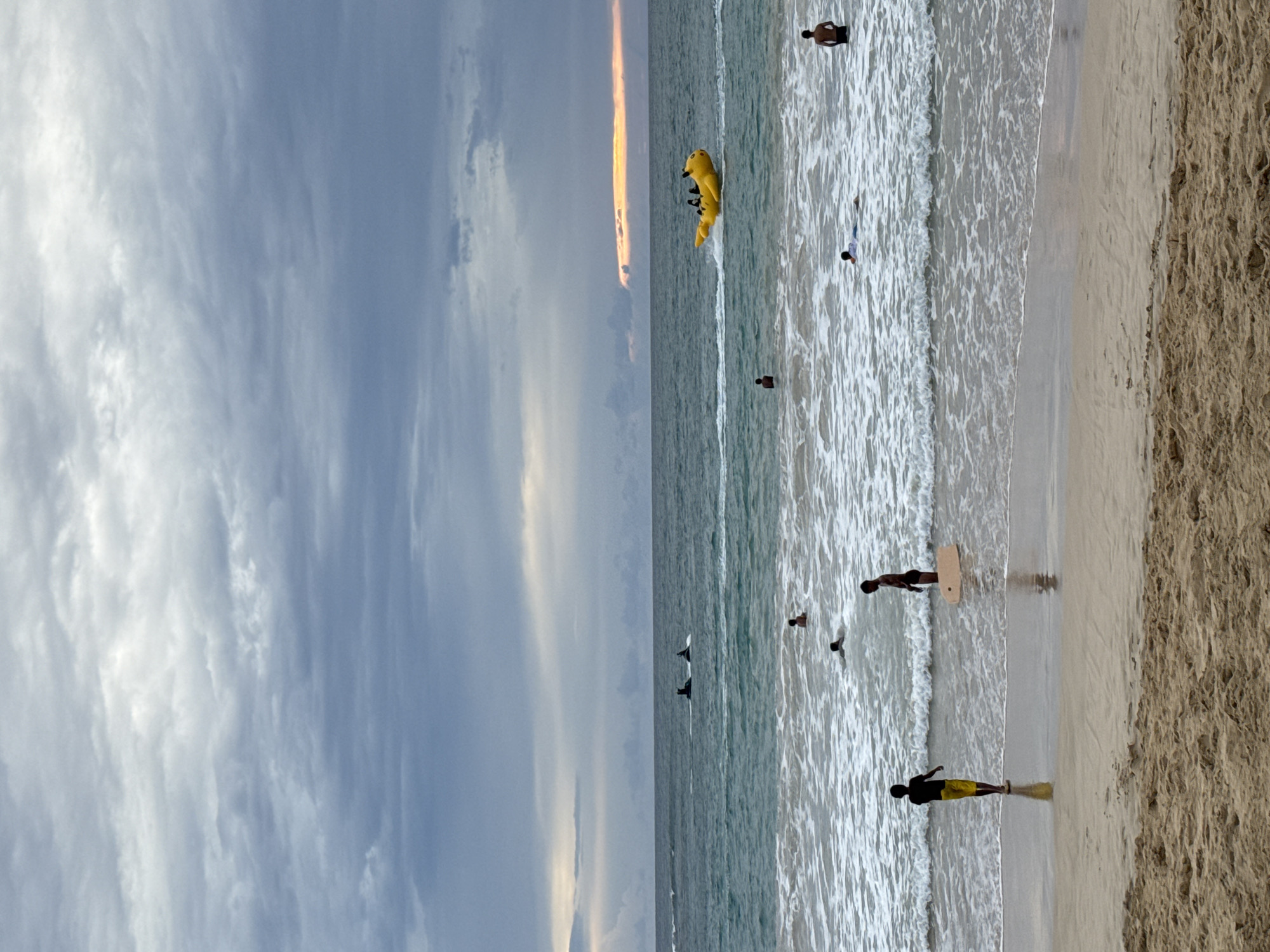 A beach scene in Meunasahbale, Indonesia, under a cloudy sky. The ocean has waves crashing onto the shore. In the foreground, two children stand on the wet sand, one wearing a black shirt and yellow shorts, the other wearing black shorts and holding a light brown surfboard. Further out in the water, several other individuals are present, some swimming and others standing. In the distance, a yellow banana boat with several passengers is being towed by a speedboat. Two jet skis are also visible further out in the water. The sand in the foreground is a light brown color. The sky is filled with heavy gray clouds, with a sliver of orange light visible on the horizon.