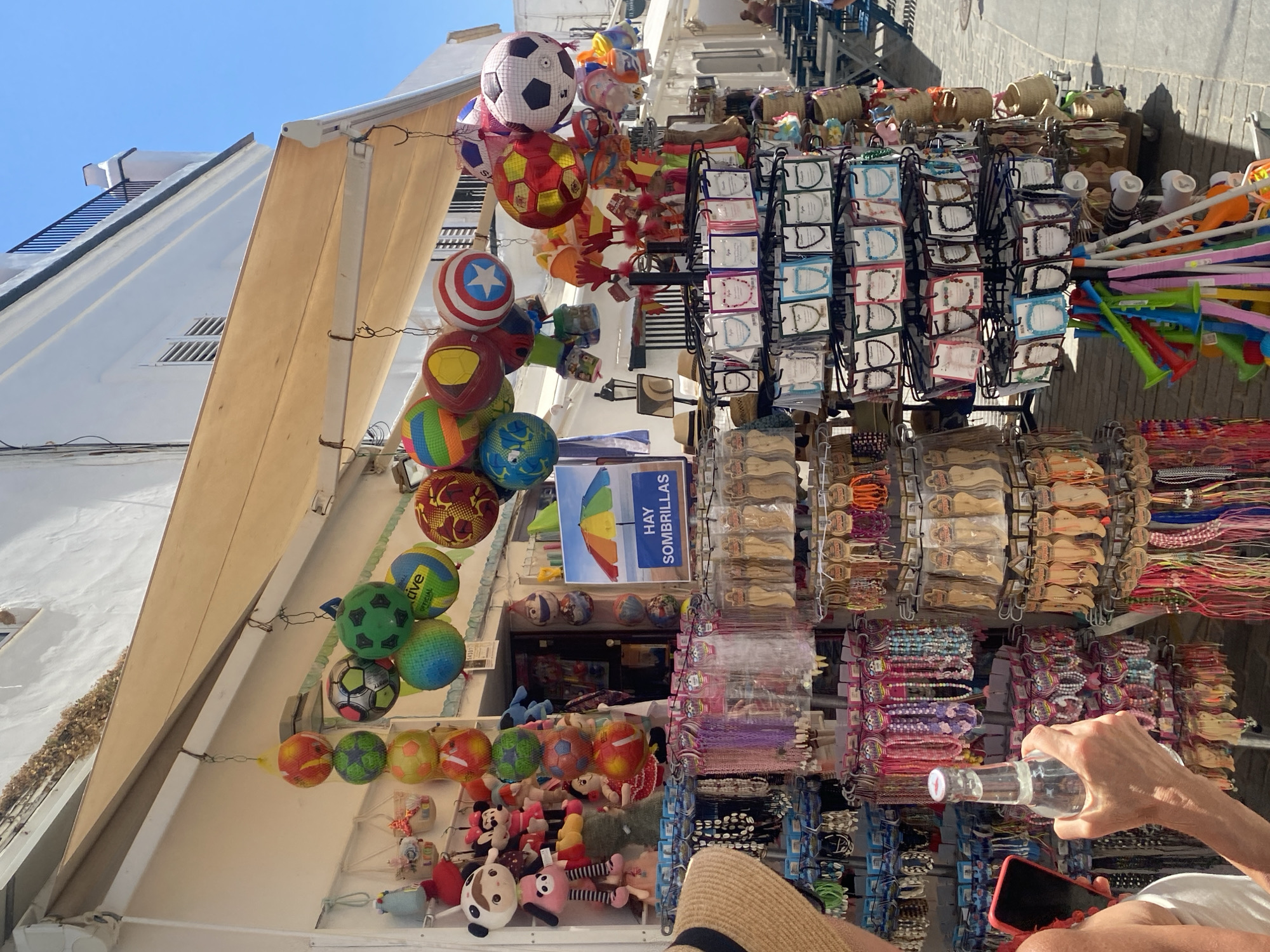 The image depicts a street scene in Conil De La Frontera, Spain, likely a shopping area. In the foreground, a person's arm and hand are visible, holding a clear glass bottle. The person is wearing a white top and a straw hat. To the left of the person, a red smartphone is partially visible.
The primary focus of the image is a stall displaying various merchandise. Hanging from the awning are several inflatable balls, some designed to look like soccer balls, others brightly colored. Below these, racks are filled with an array of items, including what appear to be necklaces, bracelets, and perhaps keychains, presented in plastic packaging. Wooden items, possibly souvenirs, are also displayed on one side of the stall. Further down the street, other stalls and tables are visible, suggesting a market or a row of shops. The architecture features white buildings and a blue sky overhead. A sign with a rainbow-colored umbrella and Spanish text ("HAY SOMBRILLAS") indicates the sale of umbrellas. The scene appears to be outdoors on a sunny day.