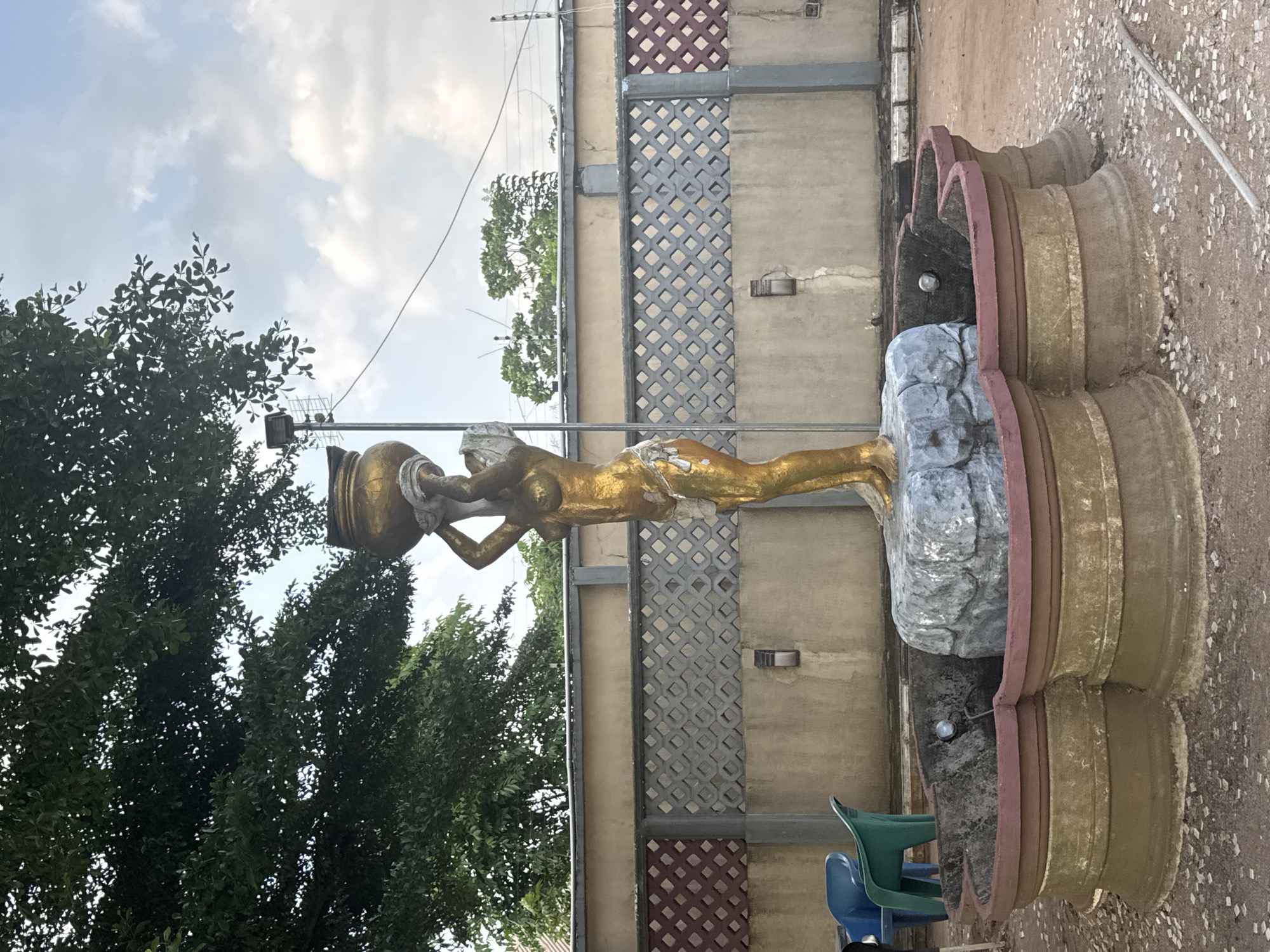 The media file displays an outdoor scene, rotated 90 degrees clockwise, featuring a prominent statue.
The central subject is a golden-colored statue of a nude woman, standing upright. She holds a large urn or jar above her head with both hands, positioned as if pouring its contents. The woman stands on a grey, rock-like pedestal. This pedestal is integrated into a larger, multi-tiered structure, likely a decorative fountain or planter, which features reddish-brown and beige/gold segments.
In the background to the left, lush green trees with dense foliage are visible, partially obscuring the sky. To the right of the statue, a building wall or fence serves as the backdrop. This wall is light brown or beige and incorporates sections of decorative lattice or grille patterns in grey and a darker reddish-brown. A small outdoor light fixture is mounted on the wall.
The ground area around the base of the structure appears to be concrete or paved, scattered with small, light-colored pebbles or debris. Two plastic chairs, one green and one blue, are situated near the statue's base. Overhead, thin wires or cables are visible, stretching across the frame, along with a small antenna-like structure.
The sky is partly cloudy, indicating daytime. No specific activity or event is taking place; the image captures a static outdoor setting. There is no visible text. The location context provided is Ihiagwa, Nigeria. The overall mood is neutral and observational.