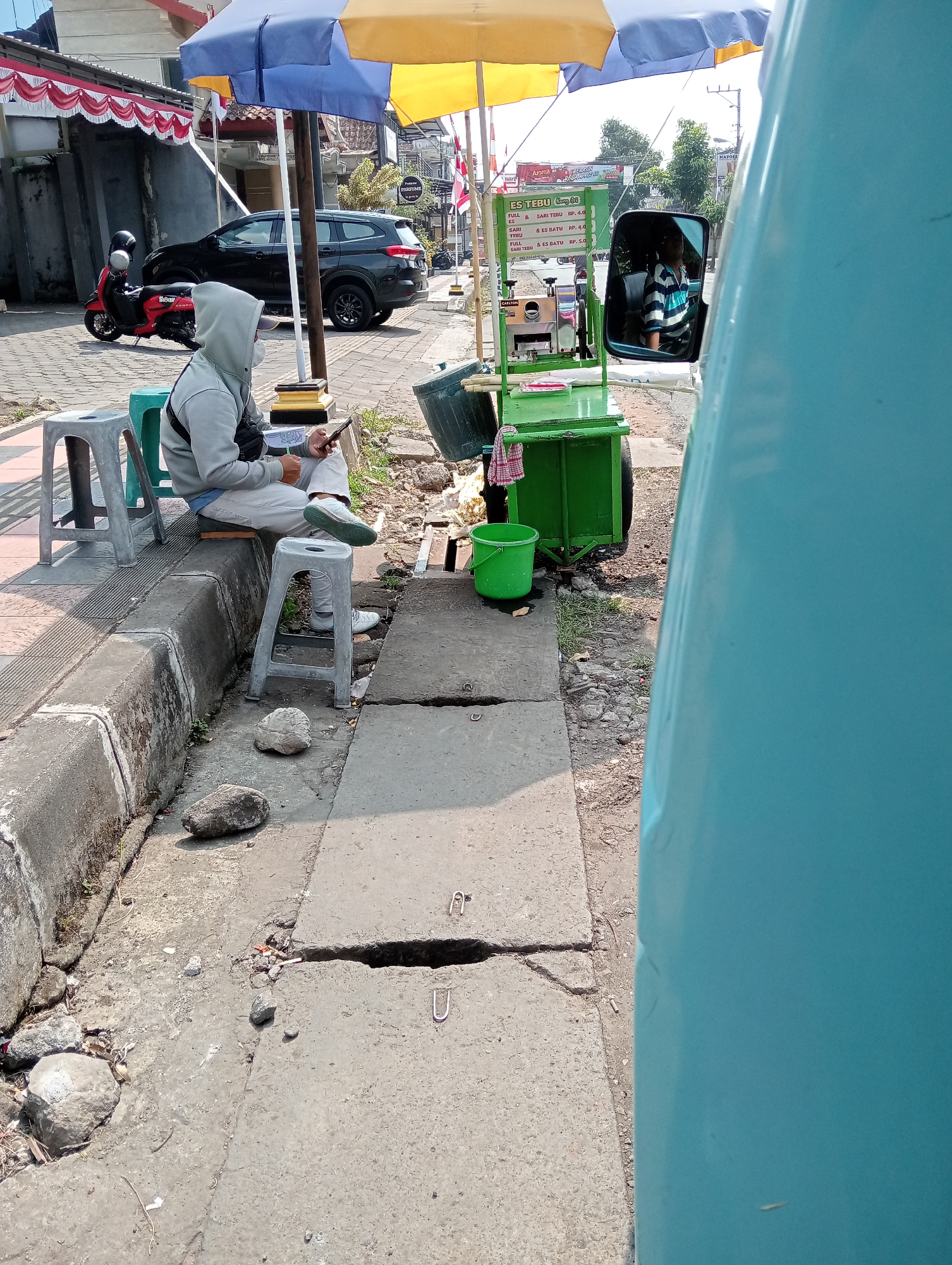 A person wearing a grey hoodie and white pants sits on a grey stool next to a green cart with a yellow and blue umbrella. The person is looking at a mobile phone and holding a pen. A red scooter is parked behind the person, and a black SUV is visible in the background. The scene appears to be a street in Grabag, Indonesia, with a sidewalk, curb, and a road. There are also several rocks and debris on the ground. The image is taken from a low angle, possibly from another vehicle.