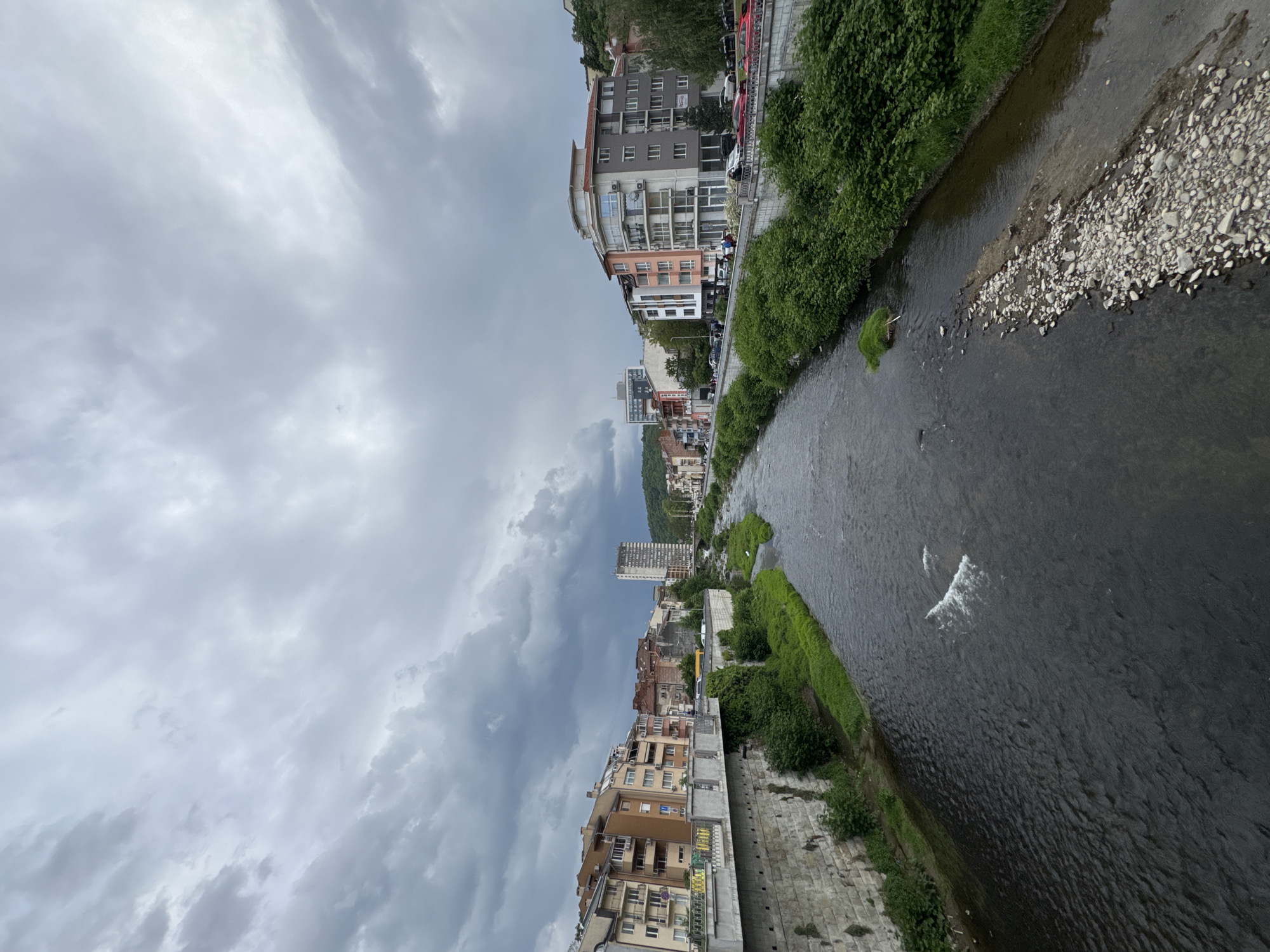 The image shows a view along the Yantra River in Gabrovo, Bulgaria. Buildings line both sides of the river, varying in height and architectural style. The river's water flows with visible ripples and patches of lighter-colored gravel and vegetation along the banks. The sky is overcast with gray, heavy clouds. There are no visible people or animals in the scene. Cars are parked along the side of the river where the buildings are located.