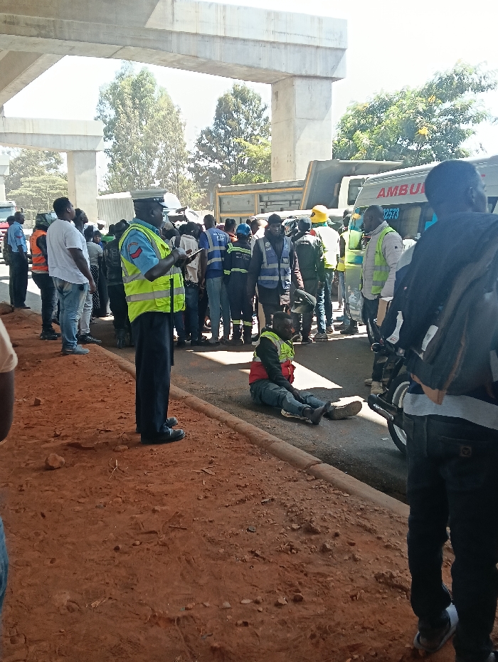 The image displays an incident scene beneath an elevated concrete highway structure, identifiable as a section of the Nairobi Expressway in Nairobi, Kenya. A uniformed male police officer, wearing a blue shirt, dark trousers, a high-visibility yellow-green vest, and a dark cap, stands in the center-left, holding and operating a mobile device. To his immediate right, a male individual in a red jacket and a yellow-green high-visibility vest is seated directly on the dark asphalt road surface, with his legs extended, adjacent to a concrete curb.
A crowd of approximately 15 to 20 individuals, mostly male, are gathered around the sitting man and the police officer. Several individuals in the crowd are wearing high-visibility vests of various colors, including blue, yellow, and green. In the background, a white ambulance with red "AMBULANCE" lettering and the number "2573" is partially visible on the right, situated behind a large, dark-colored dump truck on the left. A motorcycle's front wheel and handlebars are discernible on the right side of the frame, near the sitting individual. The foreground is composed of reddish-brown, uneven earth, bordered by a concrete curb separating it from the asphalt road. The scene is illuminated by bright daylight, with green trees visible in the distant background beyond the concrete overhead structure.
