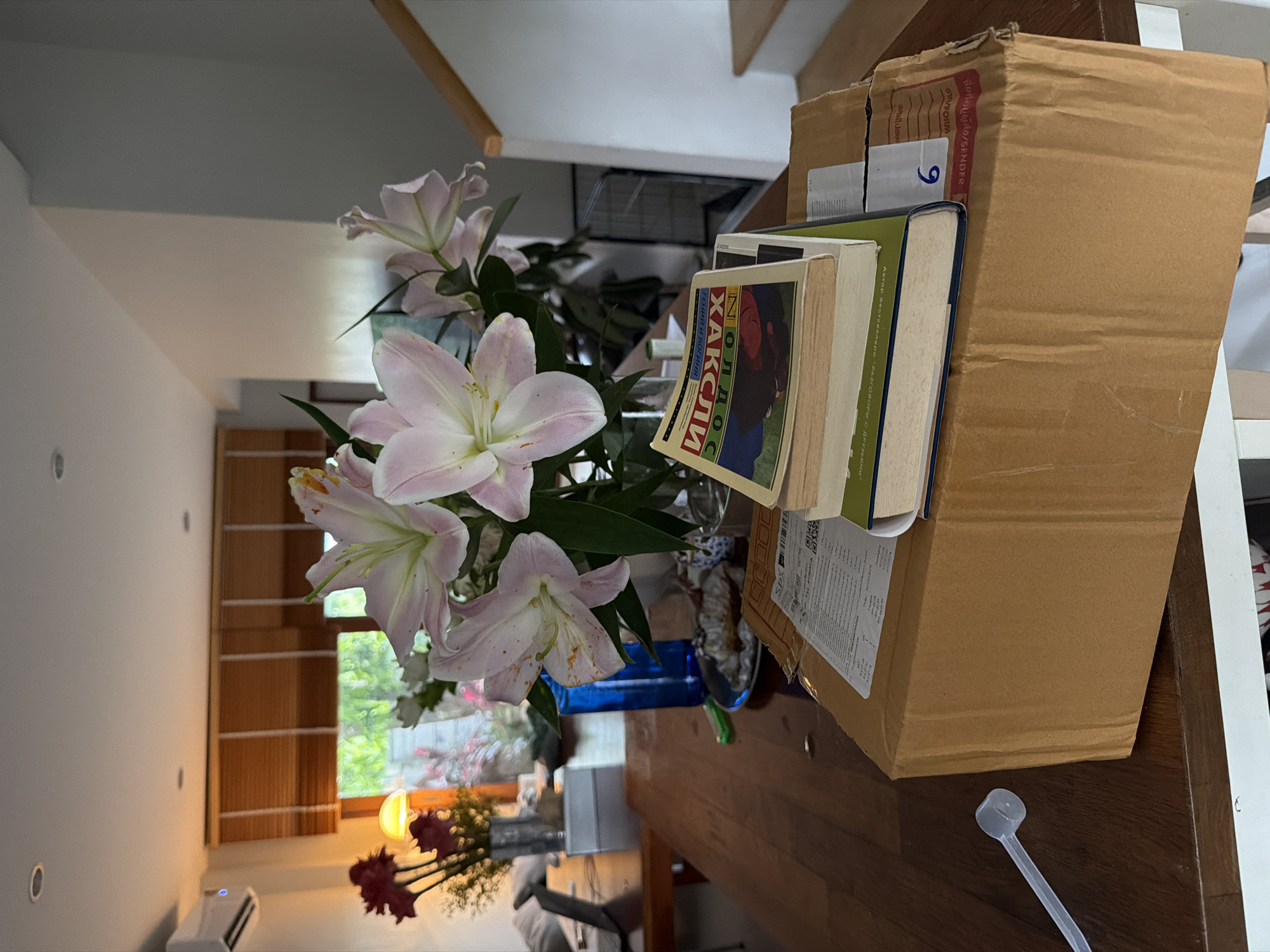 In a residential interior in Chang Phueak, Thailand, a brown cardboard box rests on a wooden table, topped by a stack of books. The uppermost book features a colorful cover with Cyrillic text. To the left of the box, a clear plastic scoop lies on the table. In the background, a vase of pale pink lilies and dark red flowers is positioned on the table. Behind the flowers, a blue glass bottle is visible. Further in the background, a window with wooden blinds looks out onto greenery. A portion of a staircase is visible on the right side of the image. The lighting suggests it is daytime, with artificial light also present.