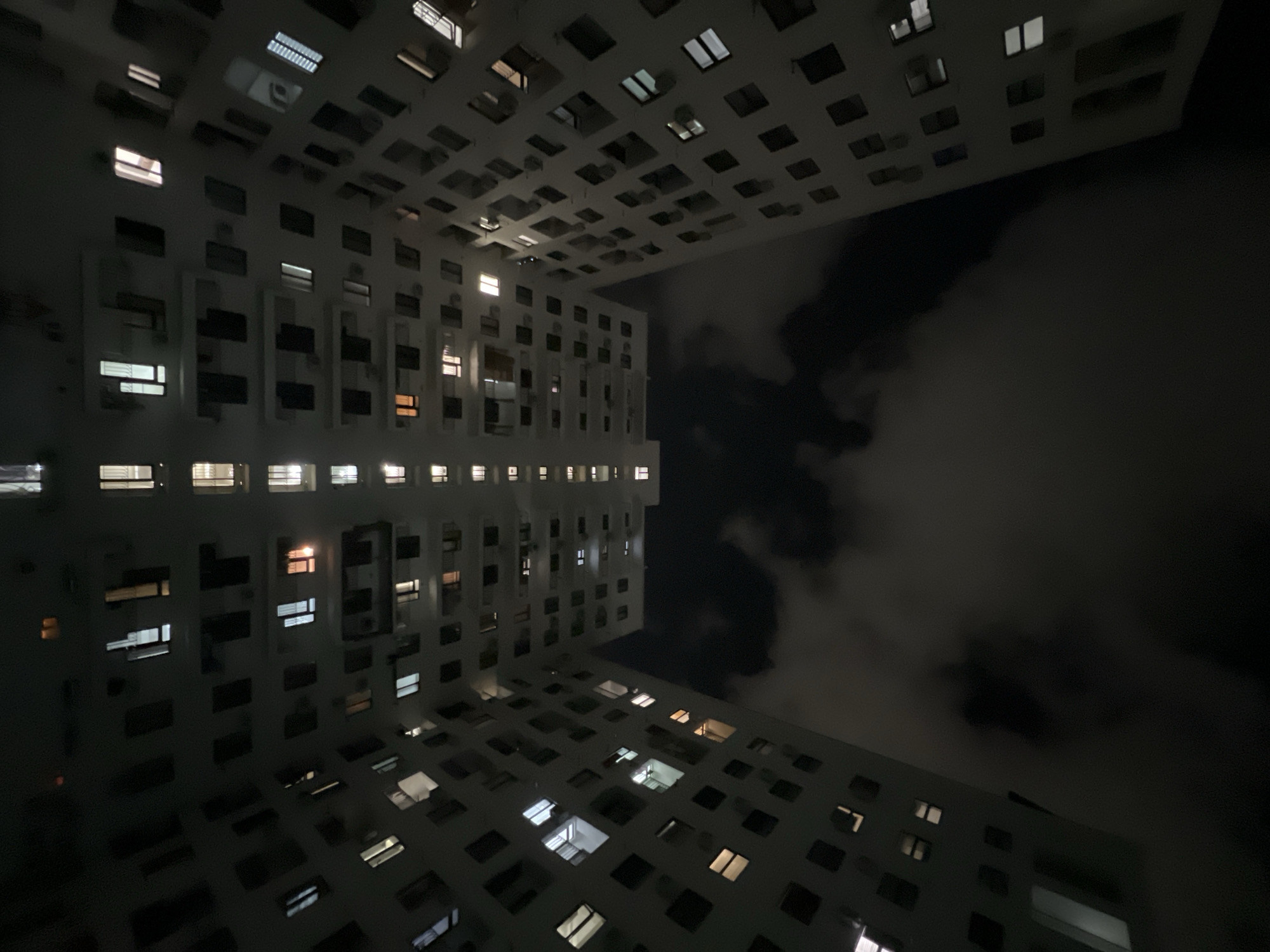 The image depicts a night scene in Yerevan, Armenia. Two high-rise buildings with numerous illuminated windows frame a dark sky filled with clouds. The buildings appear to be residential apartments. The composition suggests a low-angle view, potentially from a courtyard or street between the buildings, emphasizing their height. The lighting in the windows indicates occupancy. There are no people visible. The environment is urban at night, with the focus on the architecture and the contrast between the artificial light and the darkness of the sky.