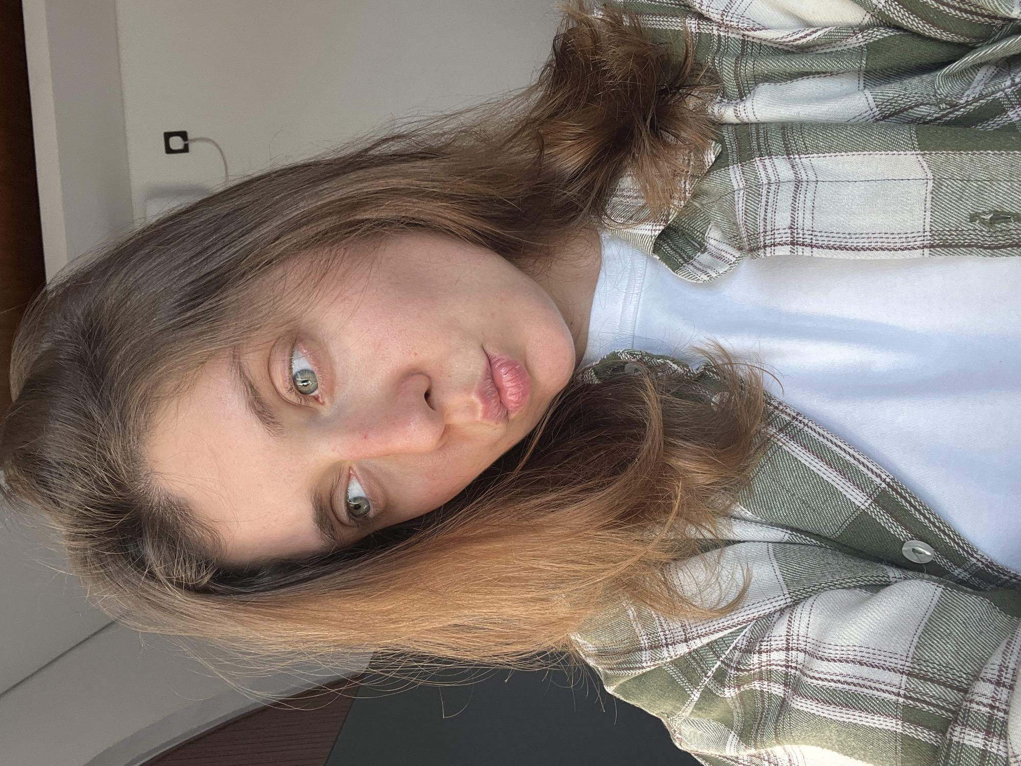 A fair-skinned person with shoulder-length brown hair and green eyes is pictured from a close, slightly angled perspective. They are wearing a white t-shirt and a partially buttoned, long-sleeved flannel shirt with a green and white plaid pattern. The background is a light grey wall, with an electrical outlet visible in the top left corner. The person's facial expression appears to be a slight pout. The environment suggests an indoor setting. It is not possible to infer the location of North Kuta, Indonesia from the visual information provided.
