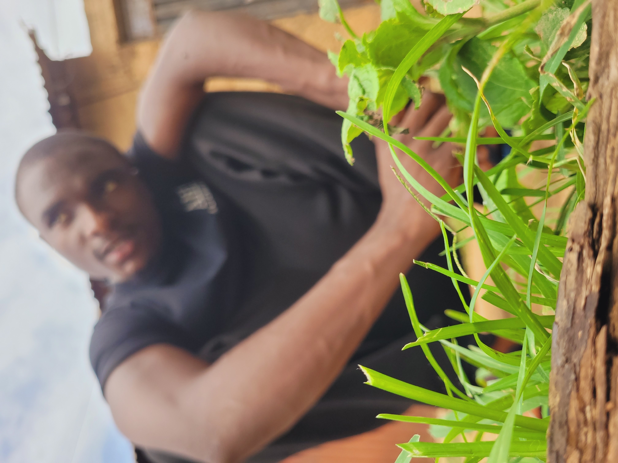A close-up, low-angle shot shows a man, out of focus, in a dark t-shirt and trousers. His arms are bent, with one raised near his head. The man is dark-skinned, likely of African descent. In the foreground, grass and green leaves are visible along with a segment of brown wood. The blurred background suggests an exterior environment, potentially in Bukuru, Nigeria. No specific landmarks or contextual cues are present to confirm the exact location.
