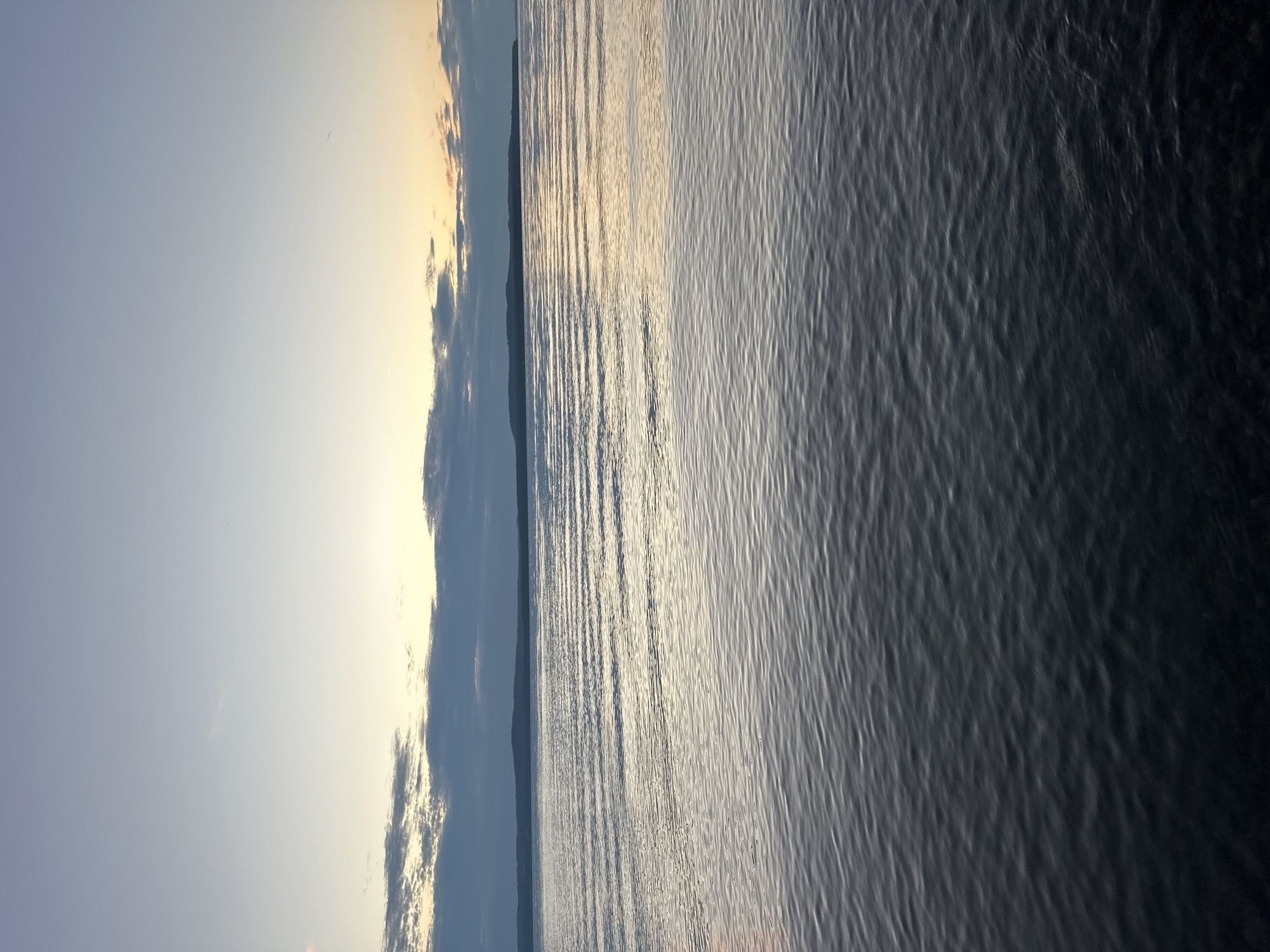 The image shows a vast body of water with gentle ripples reflecting the soft light of a twilight sky. In the distance, a low-lying landmass or island with a dark silhouette is visible on the horizon. Above the water, the sky transitions from a pale blue at the top to a warm gradient of yellow and orange near the horizon, where scattered clouds are present. The overall impression is one of tranquility and a serene natural landscape. The location is described as Горы, Russia.