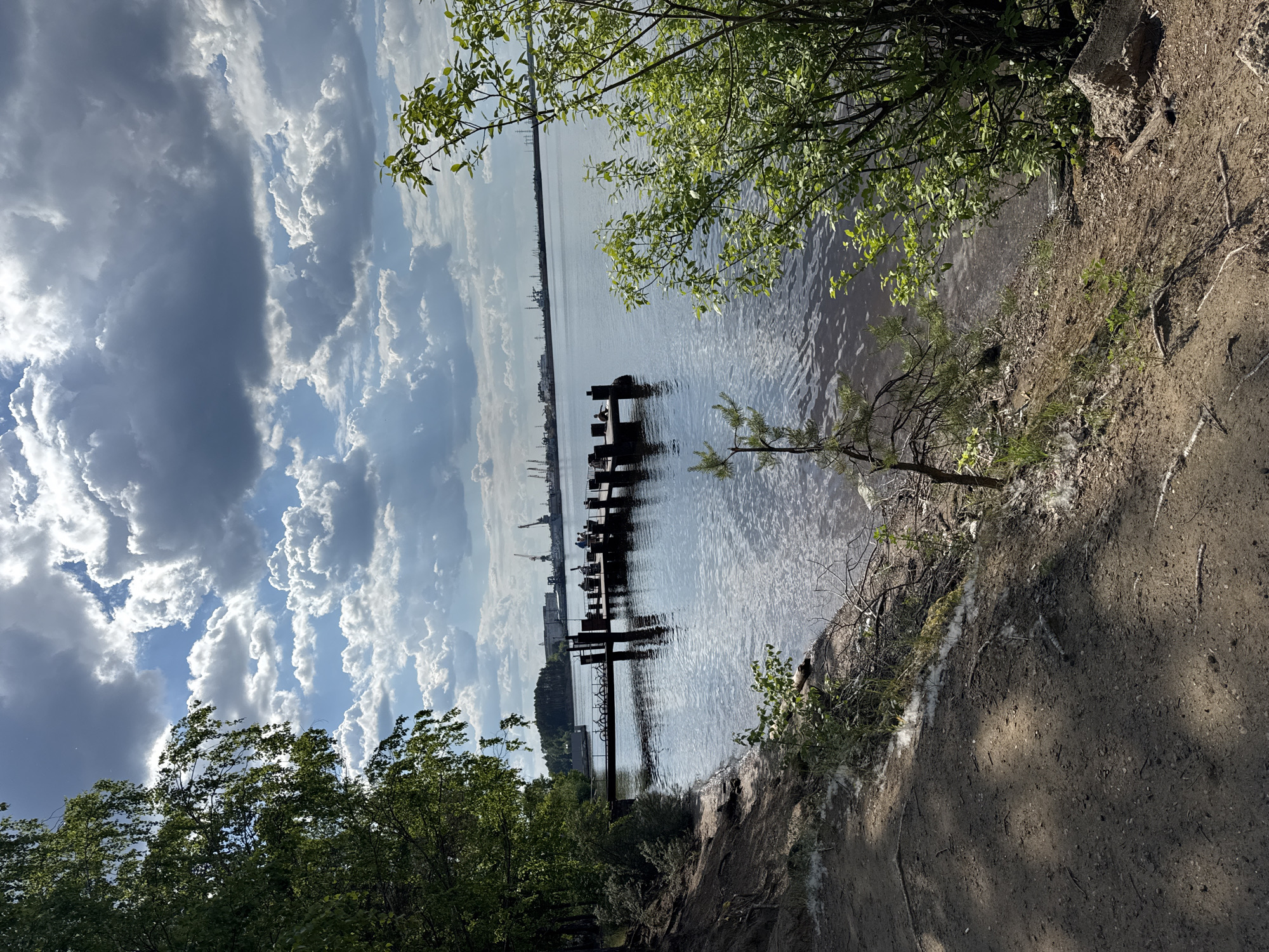 The image depicts a scenic view of the Kama River near the city of Chaikovsky, Russia. In the foreground, a sandy path runs along the riverbank, bordered by lush green trees and vegetation. The path is dappled with sunlight and shadows, suggesting a partly cloudy day.
In the middle ground, a wooden pier extends into the tranquil waters of the Kama River. Several bicycles are parked on the pier, and a few people are visible, some seated and others standing, enjoying the waterfront. The water's surface reflects the dynamic sky, which is filled with dramatic cumulus clouds.
In the background, a distant industrial landscape with cranes and structures hints at the presence of port or manufacturing facilities along the river. The overall atmosphere is serene and natural, juxtaposed with the hints of industrial activity in the distance.