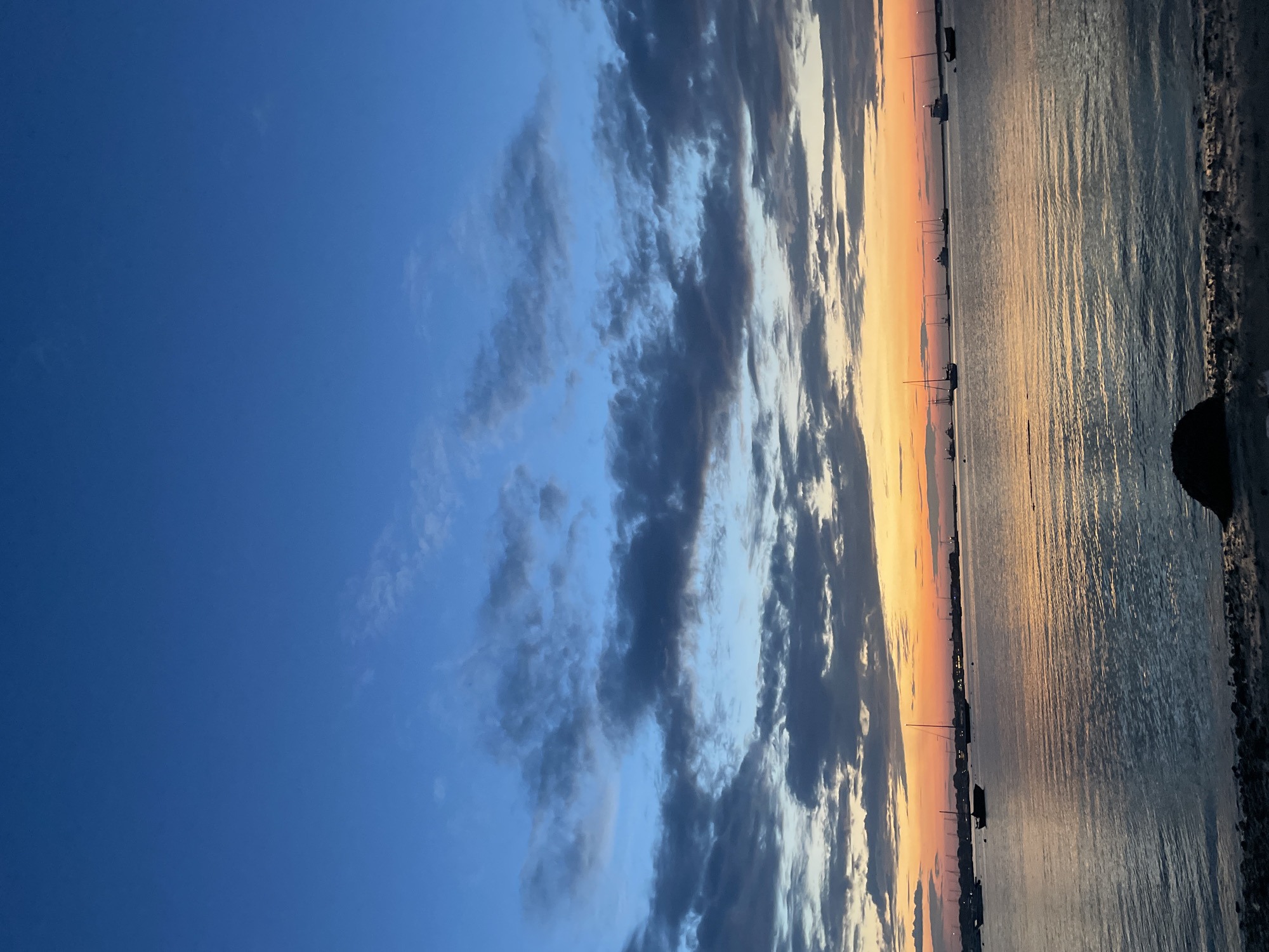 The image captures a serene sunset over the water in Nantucket, Massachusetts, United States of America. The sky transitions from a deep blue at the top to vibrant hues of orange and yellow near the horizon, with scattered clouds adding texture to the scene. Several sailboats and larger vessels are visible anchored in the calm water, silhouetted against the colorful sky. The water reflects the colors of the sunset, creating shimmering patterns. The foreground shows a pebbly shoreline with a dark, rounded rock. The overall atmosphere is tranquil and picturesque.