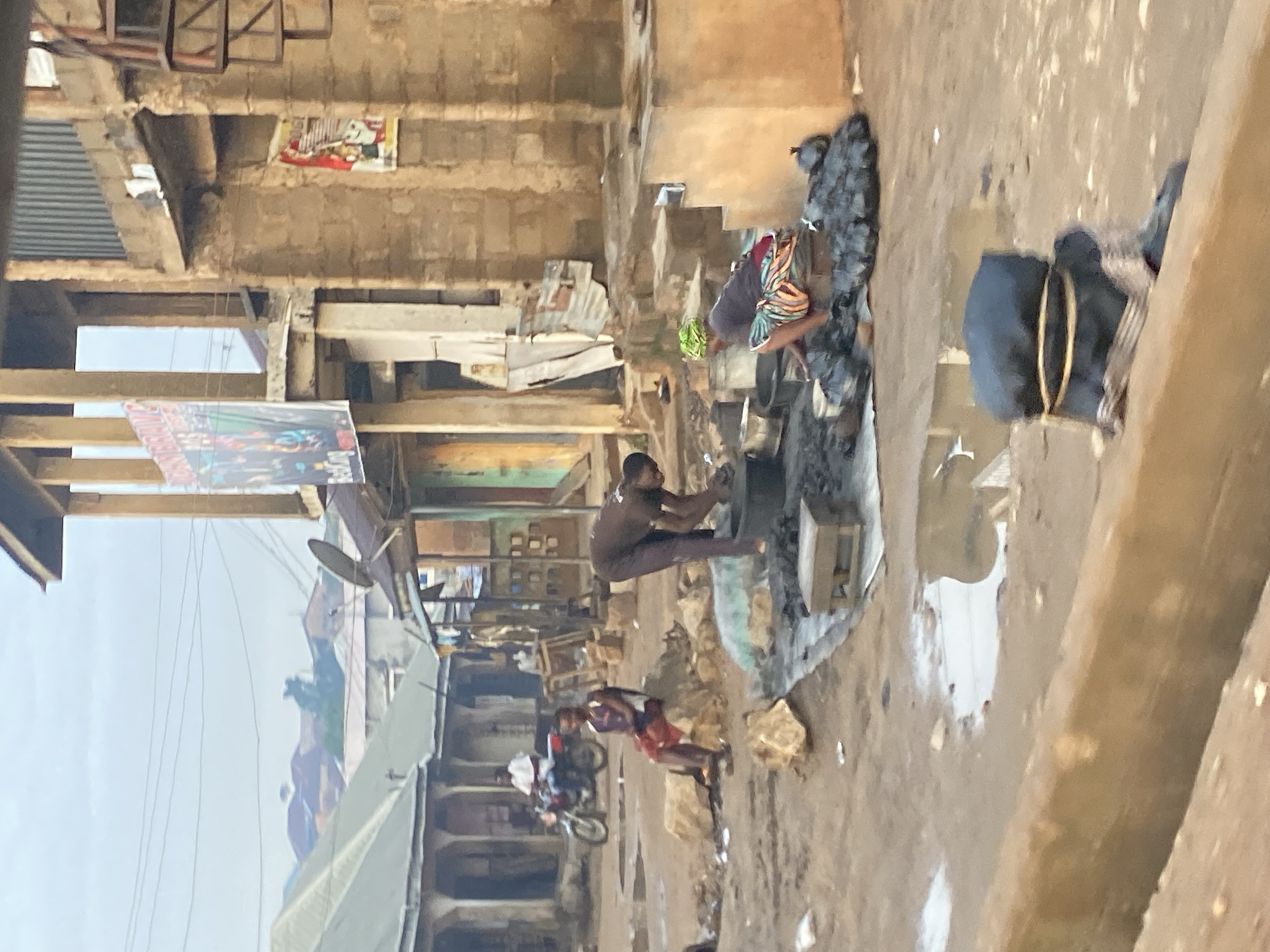 In Gwong, Nigeria, this image captures a scene within a dusty, outdoor marketplace. The sky is overcast, suggesting either early morning or late afternoon. In the mid-ground, two individuals are engaged in activity. One person, wearing a dark brown shirt and pants, is bent over, seemingly working with some materials in a large basin or pot. Another person, wearing a dark shirt and a patterned skirt and green headscarf, is also bent over, tending to items. Surrounding them are piles of black plastic bags, likely containing goods or waste. A small, wooden stool is placed nearby. In the background, a man on a red motorcycle passes by, while another man sits on a rock. Buildings with corrugated metal and concrete construction line the marketplace. The ground is uneven and damp, with puddles of water visible, indicating recent rain or poor drainage. A bundle of dark fabric, tied with rope, rests on a low, earthen wall in the foreground.