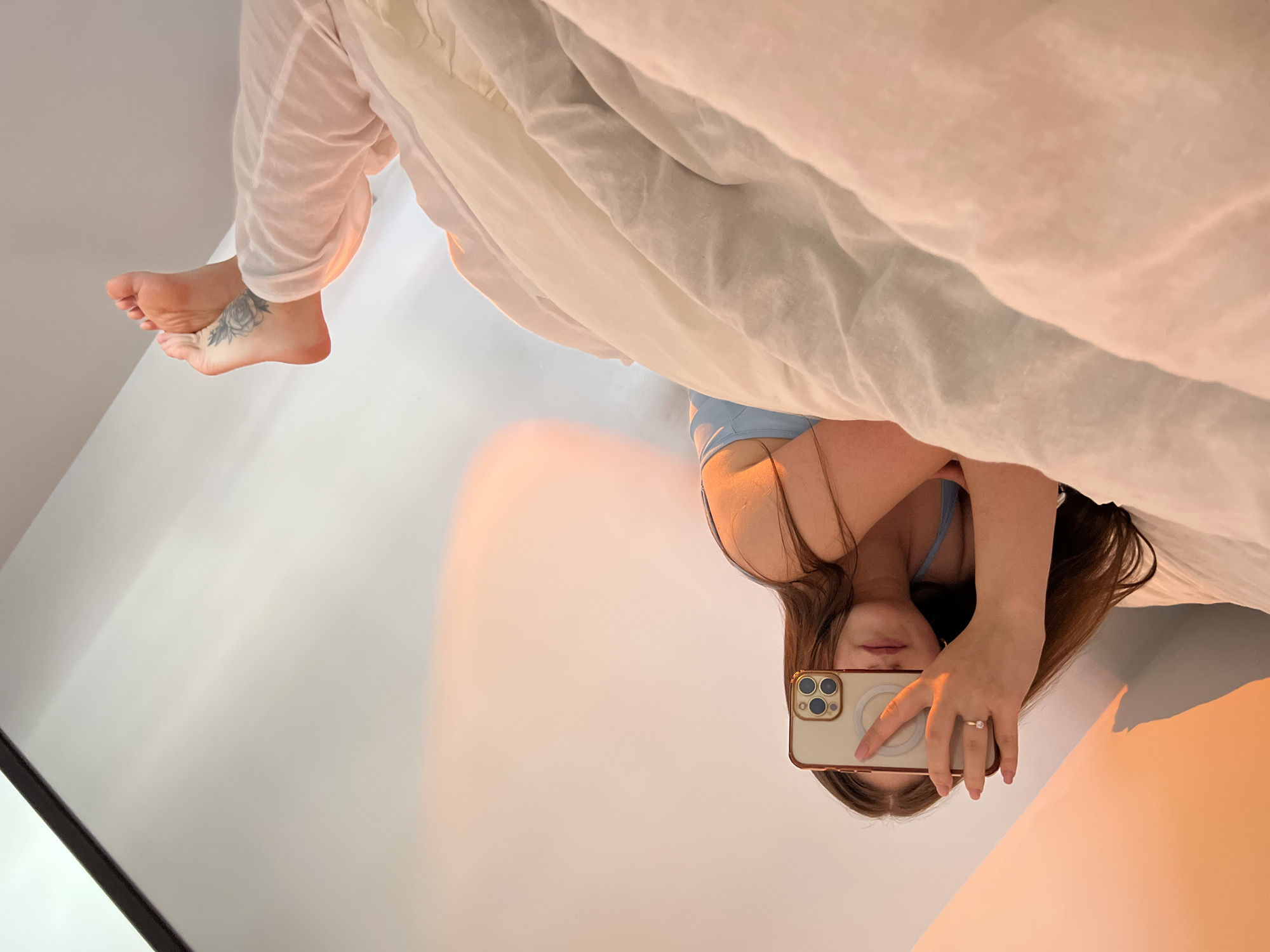 A person is taking a photo of themselves with their phone in a mirror. The person is upside down in the image, and is wearing light blue undergarments and white sheer pants. Their feet are visible with a tattoo on their left foot. Part of a white bed is visible above the person. The background is a wall painted a gradient of light and dark orange. This image does not contain information that would help determine the location in the city of Ternopil, Ukraine.