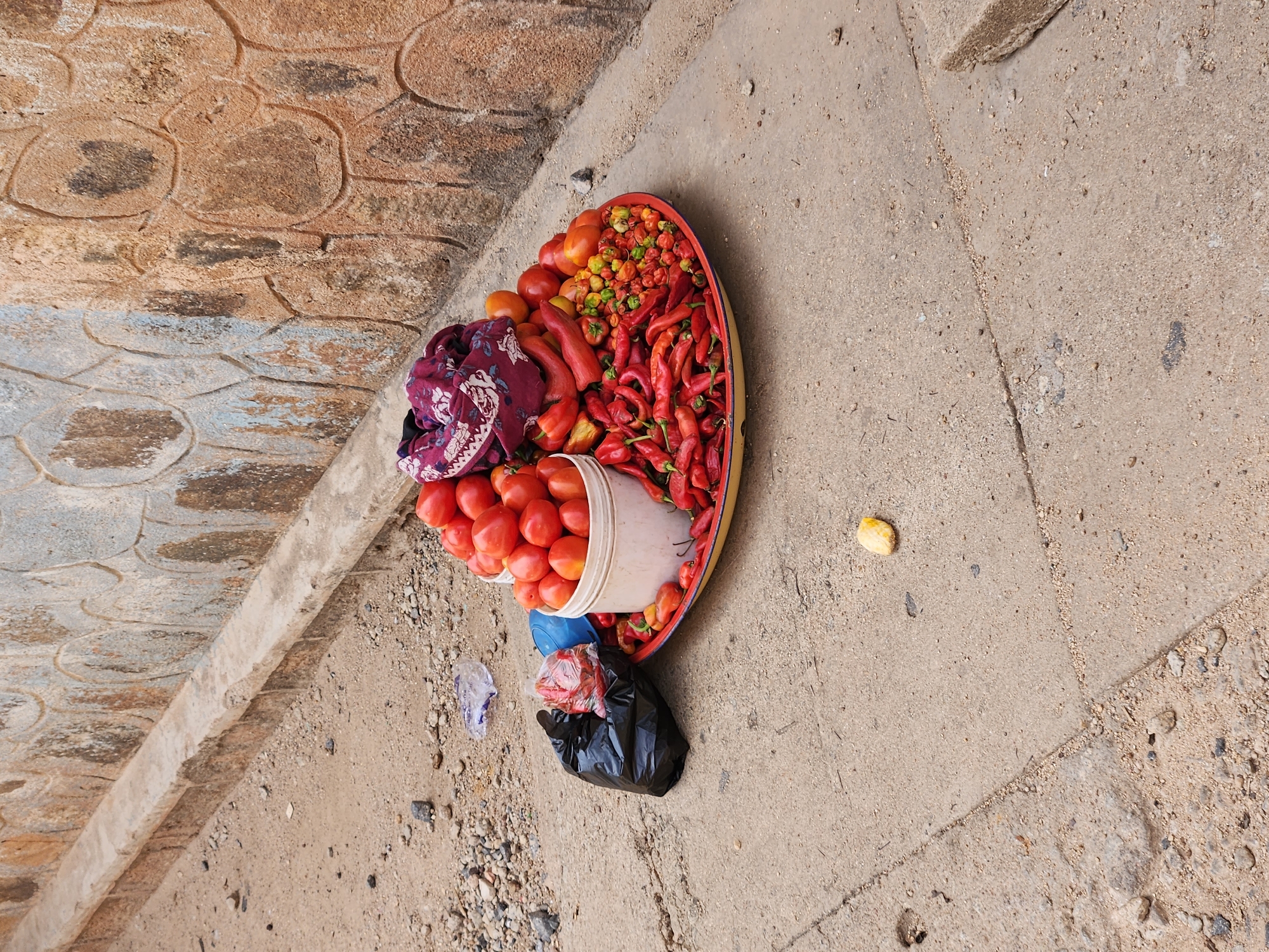 A top-down view of a street scene in Unknown, Unknown, shows a tray of produce on the ground near a sidewalk. A tray containing tomatoes, peppers (including chili peppers), and a piece of cloth sits slightly tilted on the ground. A white plastic container filled with more tomatoes is partially inside the tray. A black plastic bag and some scattered trash are near the tray. The ground consists of concrete pavement with a dividing line visible, indicating two different sections of the pavement. In the upper left of the image is a section of a stone sidewalk with a curb. A single piece of what appears to be food is visible on the concrete pavement to the side of the tray.