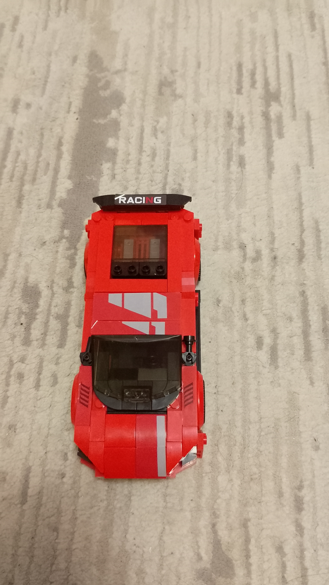 The image displays a red toy car, resembling a sports or racing vehicle, constructed from building blocks (Lego-style), positioned on a light-colored carpet. The car is viewed from a top-down perspective, occupying the lower central portion of the frame. Its primary color is bright red, accented with black elements such as a windshield, side grilles, and a rear spoiler. The spoiler prominently displays the white text "RACING." Grey stripes and abstract decals are visible on the car's hood and mid-section. A transparent, dark-tinted window area on the top surface reveals internal grey and black components. The carpet, serving as the background, is a neutral light grey with irregular, darker grey patterns, suggesting wear or a deliberate design. No people are present, and no activity is occurring. The lighting appears consistent with an indoor environment.