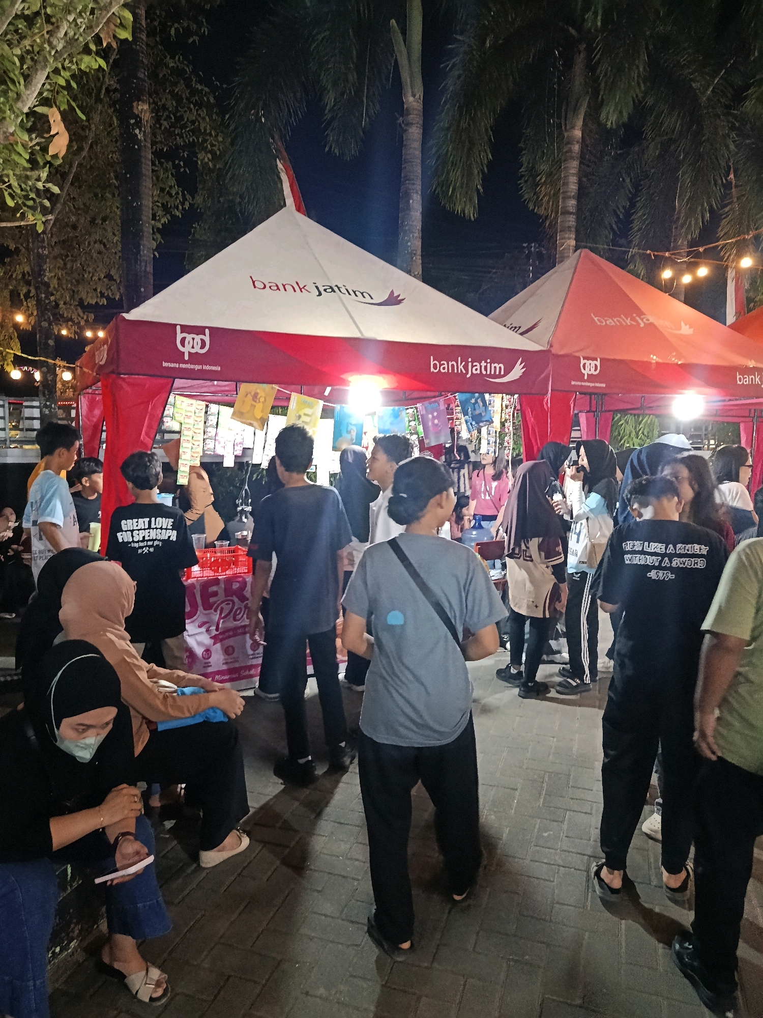 This image captures an evening scene at a market or event in Pacitan, Indonesia. Several tents are set up, two of which prominently display the "bank jatim" logo. The tents are adorned with red and white branding and seem to be selling various goods, indicated by items displayed on shelves within the tents. Palm trees and string lights are visible in the background, suggesting an outdoor setting. A diverse group of people, including men, women, and young individuals, are gathered around the tents. Some are looking at the merchandise, while others appear to be conversing. One woman in the foreground is seated, wearing a mask and looking at her phone. Another person wears a black t-shirt with the slogan "GREAT LOVE FOR SPENSA." The ground is paved with interlocking bricks. The overall atmosphere appears to be lively and social, with people engaged in shopping and interaction under the evening sky.