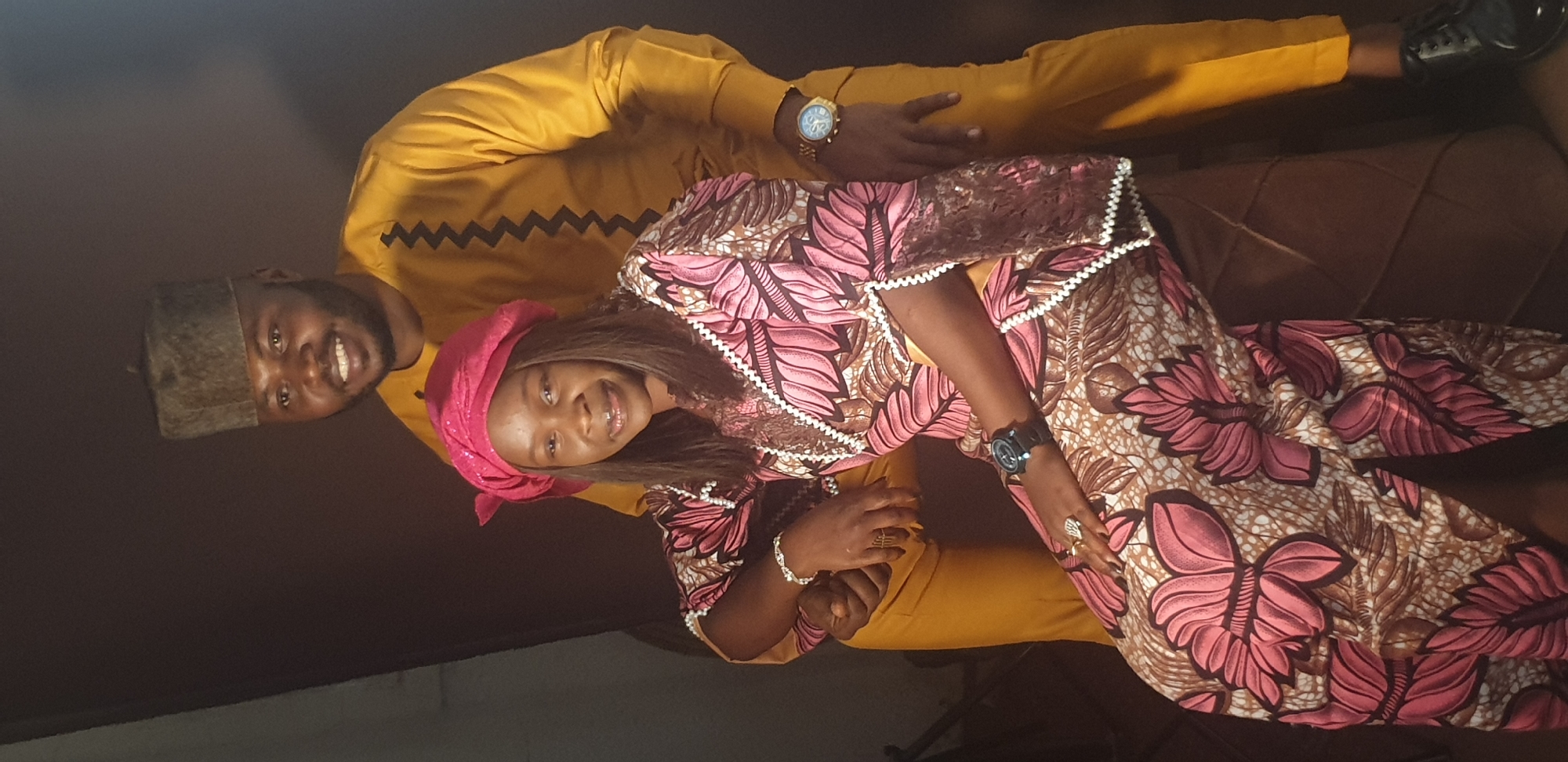 A portrait-style photograph, taken in Vom, Nigeria, features two individuals, a man and a woman, posing together and smiling warmly. The image is oriented sideways.
The man stands behind the woman, looking directly at the camera with a broad smile. He has dark skin and a short beard, wearing a mustard yellow, long-sleeved traditional top adorned with a black zigzag pattern across the chest. He also wears mustard-colored trousers and dark shoes. A brownish-grey traditional cap sits on his head, and a gold-colored wristwatch is visible on his left wrist.
The woman is positioned slightly in front of and to the side of the man, also looking at the camera with a bright smile. She has dark skin and long dark hair, partially covered by a vibrant pink headscarf. She is dressed in a long, patterned gown featuring pink, brown, and white floral motifs, with white zigzag trim along the sleeves. A silver-colored bracelet is on her right wrist, and she wears a dark-faced wristwatch on her left wrist, along with a ring on her left ring finger.
Both individuals exude happiness and affection, suggesting a joyful occasion or a cherished moment. The background is dark and indistinct, focusing attention entirely on the smiling subjects. No text is visible in the image.