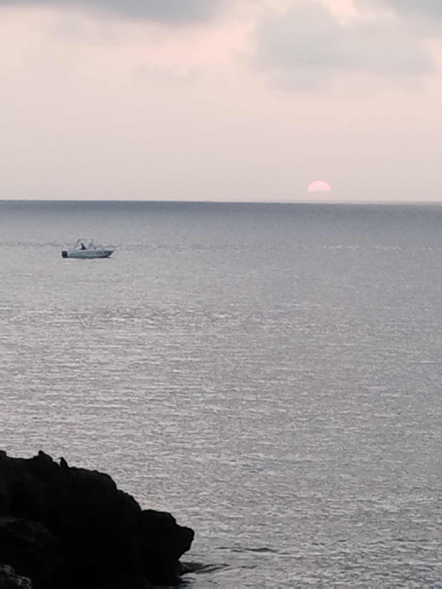 The image depicts a calm sea at dusk with a hazy, pale pink sun setting near the horizon. A small white boat with an individual visible at the helm is sailing on the water. In the foreground, a dark, rocky shoreline is visible. The scene appears to be the coast of Antibes, France, likely taken from a vantage point overlooking the Mediterranean Sea.