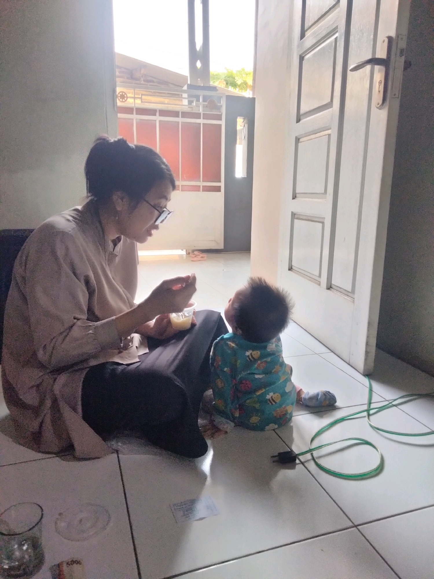 In a residential interior, possibly in Suka Sari, Indonesia, a young woman with dark hair pulled back and wearing glasses and a light-colored shirt is sitting on the floor. She is holding a small cup with yellow food in it and a utensil, appearing to be feeding a baby. The baby, seen from the back and wearing a blue outfit with bee and flower patterns, is sitting on the floor, looking upwards. The floor is tiled with light-colored squares. To the right, a white door with paneled sections is ajar, revealing a glimpse of the outdoors or another room. A green electrical cord with a black plug lies on the floor in front of the baby. Other objects on the floor include a glass, a small white disc, and a piece of paper with text, possibly currency. The lighting suggests it is daytime, with light streaming in from the open doorway and a window in the background, which has a barred metal gate.