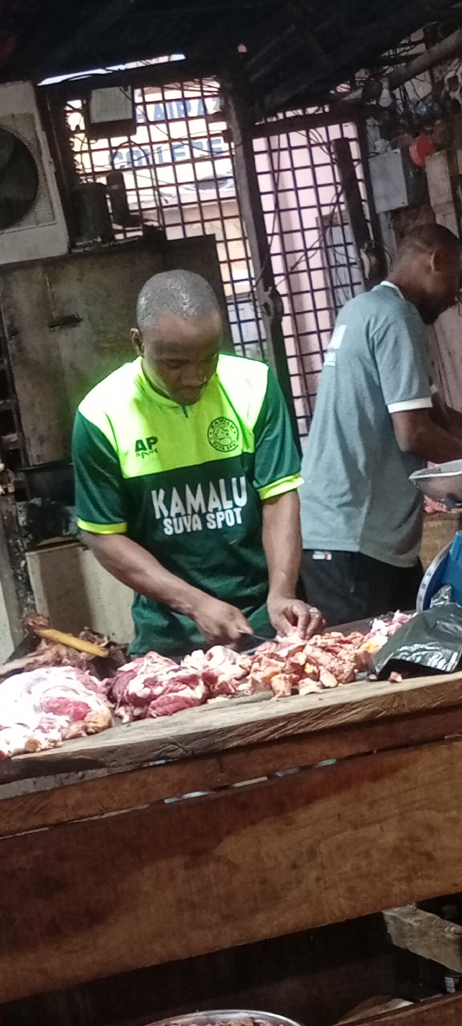 The image captures a scene inside a meat market, likely in Dorayi, Nigeria, given the writing on the shirt of the man. A man wearing a green and yellow t-shirt with "KAMALU SUYA SPOT" printed on it is seen cutting up raw meat on a wooden table. Another man in a gray t-shirt is standing behind him, also working. The environment seems to be a typical market stall, possibly with an open-air or semi-covered setting, as suggested by the barred structure in the background. There are piles of raw meat on the table, which appears well-worn. The lighting is dim, indicating an indoor or shaded location. An air conditioning unit is visible near the ceiling.
