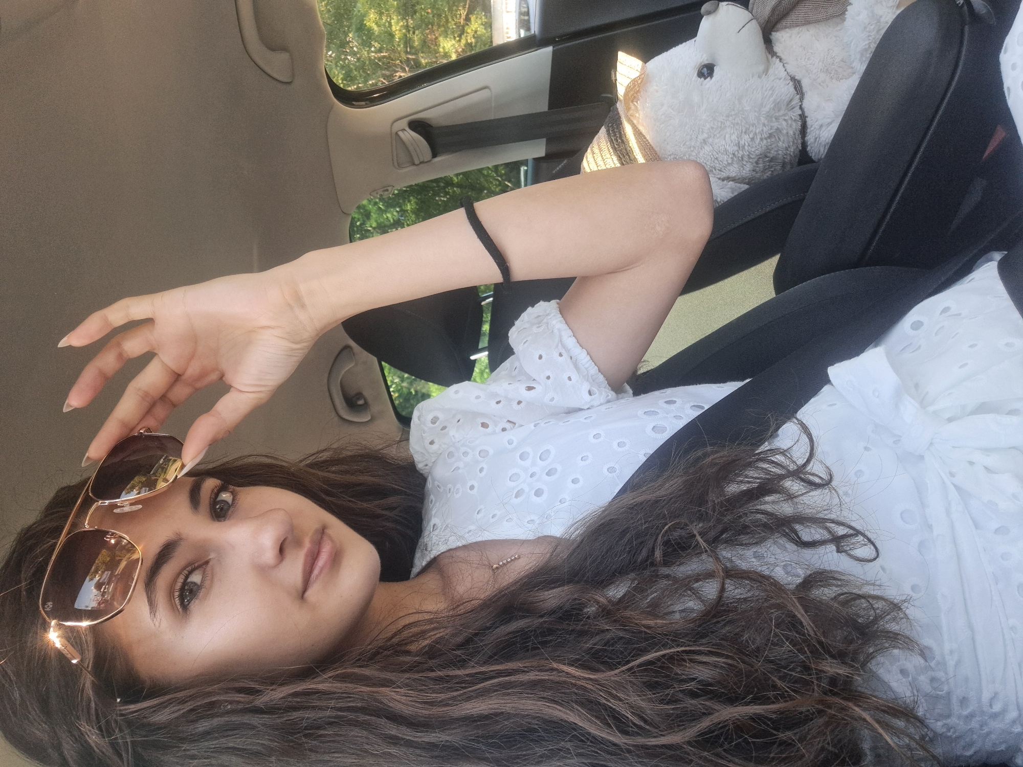 The image shows a young woman with long, dark, wavy hair, wearing a white eyelet top and sunglasses. She is in the passenger seat of a car, with her right hand raised to adjust her sunglasses. A plush white bear wearing a straw hat and scarf is visible in the background, on the passenger seat. The car interior, including a seatbelt and the car's headliner, is also visible. Outside the car, trees and foliage can be seen, suggesting an outdoor setting, possibly a drive through a park or a scenic route. The image has a warm, sunny feel, indicated by the lighting and the reflection in the sunglasses. The location is not specified to be in Sofia, Bulgaria.