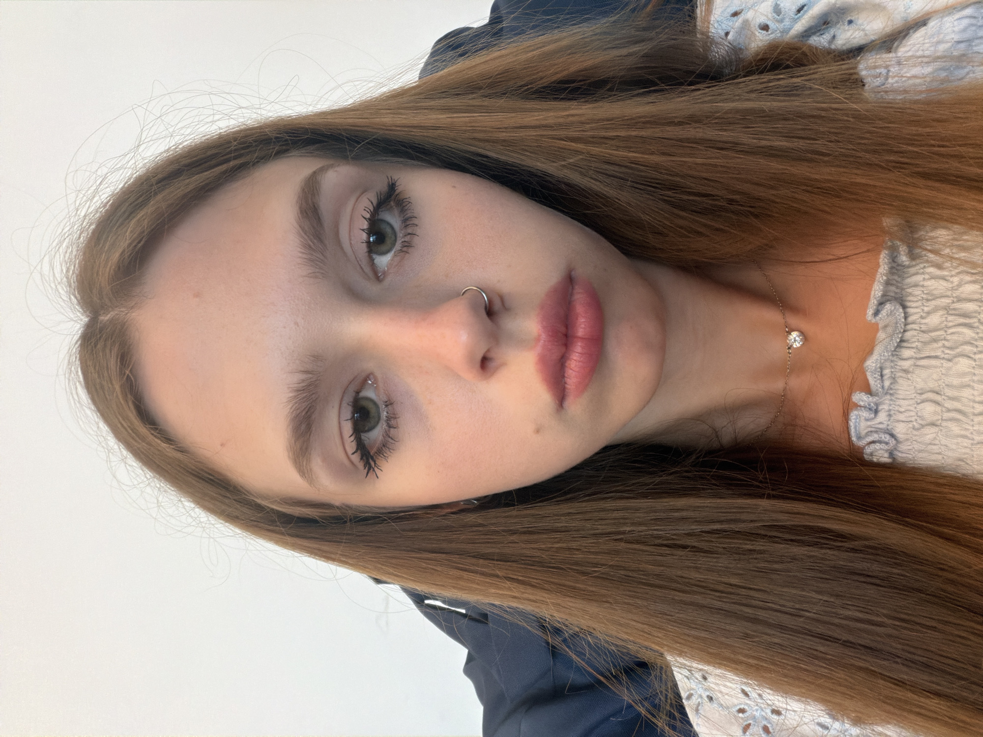 This is a close-up, portrait-style media file featuring a young person with long, brown hair. The subject is positioned at an angle, with their face tilted towards the camera, creating an intimate and personal feel. Their gaze is directed forward, though slightly off-camera, and their expression appears neutral.
Key features visible include the subject's eyes, which are open and show green irises. Their eyelashes are noticeably enhanced with mascara, and their eyebrows are well-defined. A silver hoop nose ring is visible on the subject's left nostril. Their lips are full and appear to be colored with a matte lipstick.
The subject is wearing a light blue, off-the-shoulder top with a textured, gathered fabric around the neckline. A delicate silver necklace with a small, clear pendant is visible resting on their collarbone. The background is plain and light-colored, suggesting an indoor setting, possibly a studio or a room with ample lighting. The lighting appears to be bright and diffused, casting soft shadows.
The overall impression is one of a casual, posed photograph, likely taken by the subject themselves. No specific time of day or weather conditions can be deduced from the image. There is no visible text in the media file.