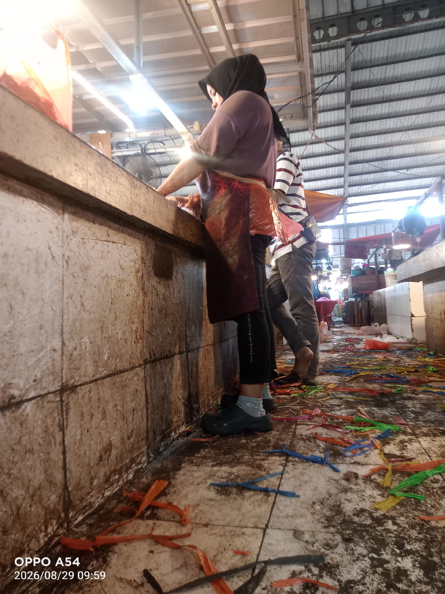 The image depicts the interior of a market in Jambi, Indonesia, as indicated by the timestamp "2026/08/29 09:59" and the style of the market stalls and wares. The scene is lit by bright overhead lights, revealing a bustling environment. In the foreground, a woman wearing a black hijab and a maroon apron is seen from the side, appearing to be engaged in work at a butcher's counter. Her hands are not visible, but her posture suggests she is handling food or products. To her right, another individual, partially visible and wearing a striped shirt and grey pants, stands with their back to the camera, also at the counter. The floor of the market is littered with colorful plastic strips, suggesting a recent event or ongoing cleanup. The market stalls are constructed of concrete and wood, with metal roofing overhead. Other parts of the market, including more stalls and stacked items, are visible in the background. The overall impression is of a busy, functional marketplace.