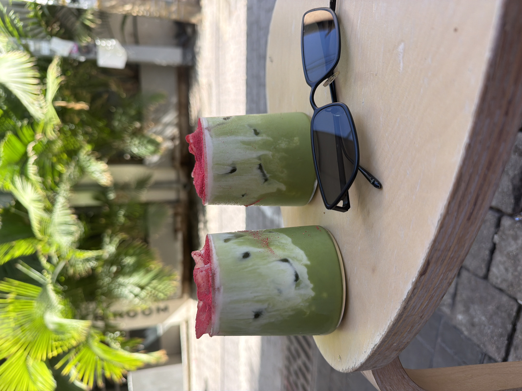 Two glasses of matcha latte with a swirl of milk and topped with pink powder sit on a light-colored, round wooden table. A pair of black sunglasses rests on the table in front of the glasses. In the background, lush green palm fronds are visible against a blurred urban setting, suggesting a cafe or outdoor seating area. The presence of palm trees and the overall ambiance are characteristic of Tel Aviv, Israel. The cafe's name, "MOOM", is faintly visible on a structure in the background.