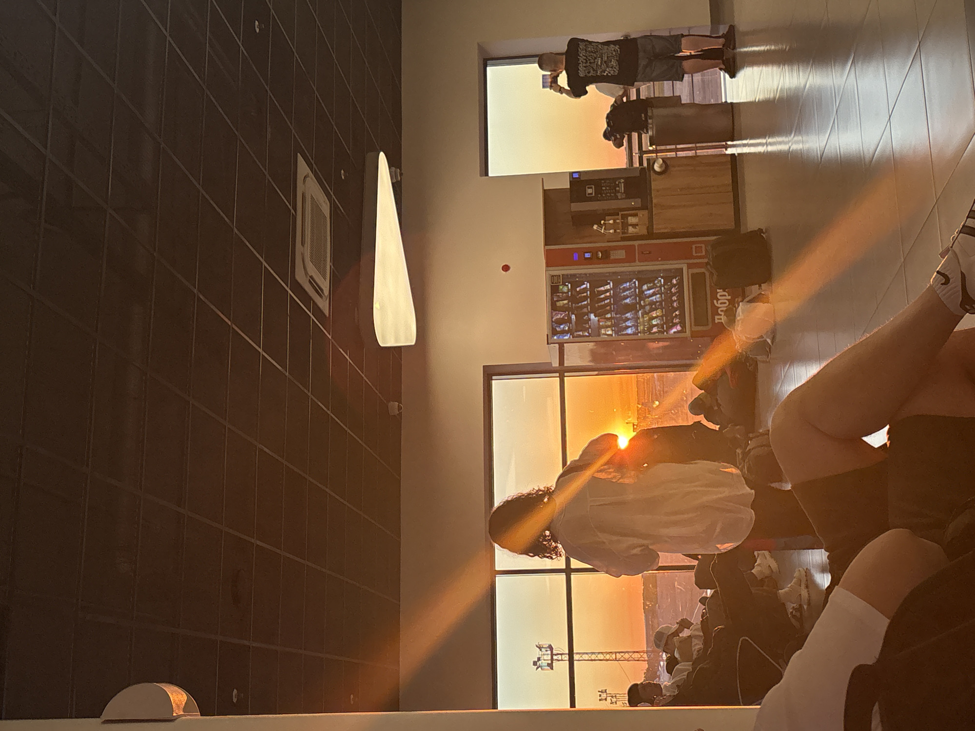 The image depicts an airport waiting area during sunset, with the low sun casting strong orange rays across the room. Several people are seated, some looking out the large window at the exterior, where airport infrastructure like towers and lights are visible against the bright sky. One person, silhouetted by the sun, is standing near the window, facing away from the camera. To the right, another individual is standing and taking a photo with a mobile phone. A vending machine is visible against the wall. The ceiling features a grid of dark tiles and a modern air conditioning unit. The floor is tiled and reflects the light. The scene suggests a moment of waiting, with passengers observing the sunset.