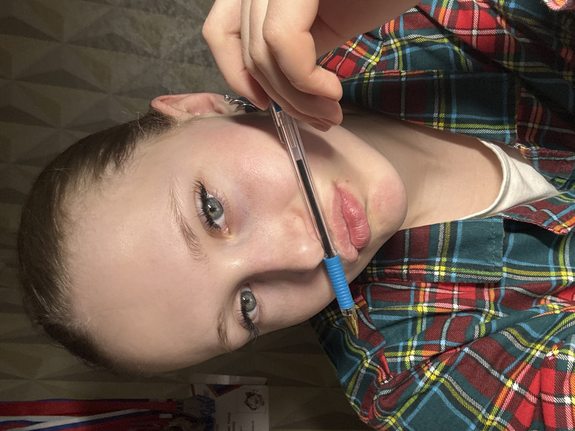A young, light-skinned person with blue eyes and light brown hair is shown from the chest up. They are holding a blue ballpoint pen vertically against their face, between their nose and upper lip. The person is wearing a plaid shirt with colors of red, green, blue, yellow, and white. A small silver earring is visible in their left ear. The background is out of focus but shows a brown surface. A small package with the Russian flag is visible on the surface in the background. There is no evidence to support a location in the city of Chaikovsky, Russia.