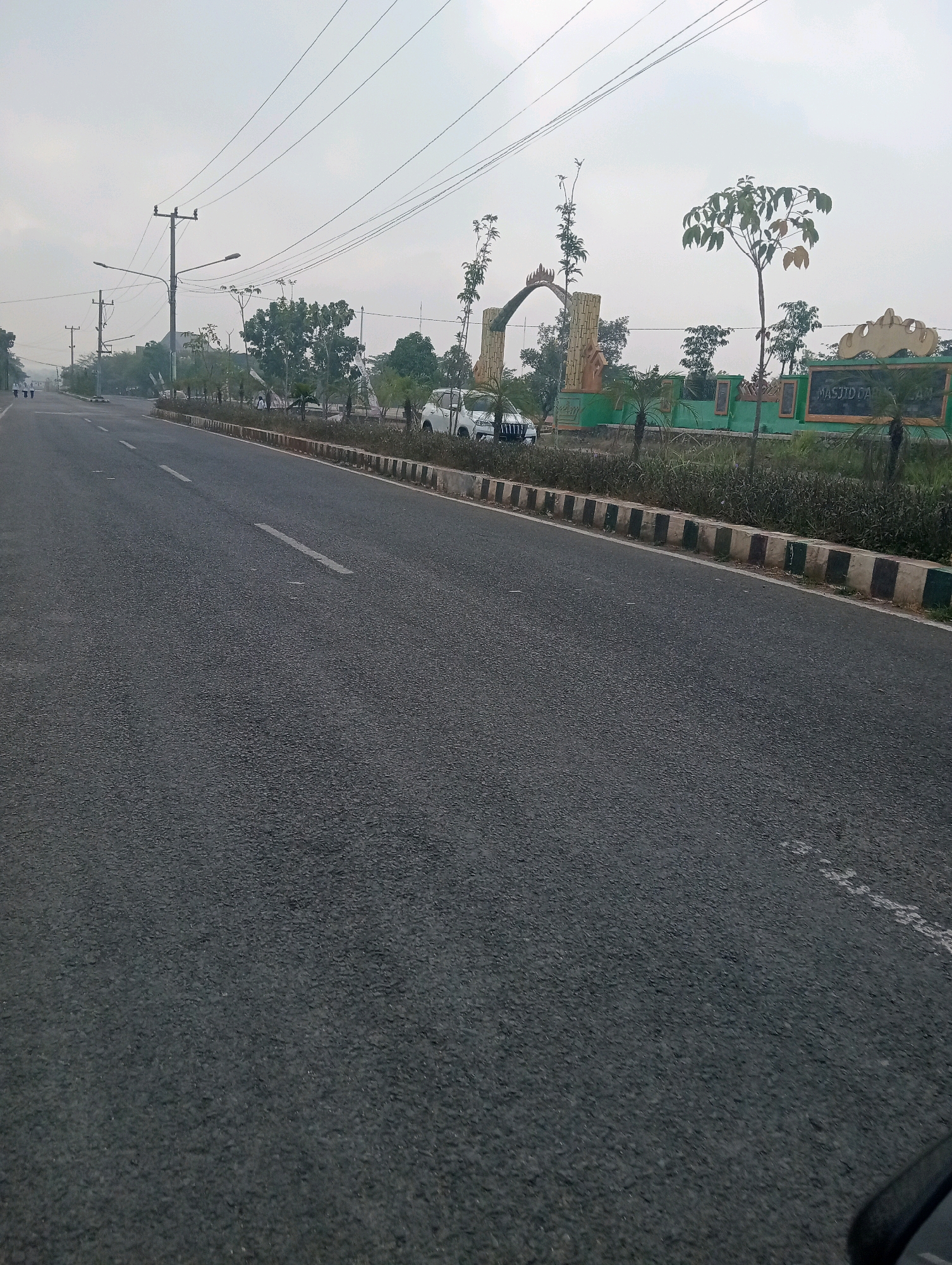 The image shows a view of a road in Sidoharjo, Indonesia. The road is paved and has white lane markings. There are several utility poles with power lines running overhead. On the right side of the road, there is a green fence with an archway and a sign that reads "MASJID DA'WATUL IMAN." Several trees and some vegetation are visible along the roadside. A white car is parked near the archway. The sky is overcast, suggesting a cloudy day. The overall scene depicts a semi-urban or suburban environment with a focus on infrastructure and a nearby mosque.