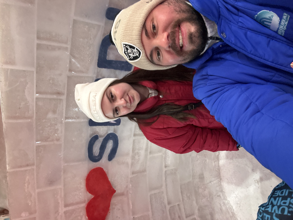 A selfie-style portrait shows two individuals inside an ice structure, identified as Ski Dubai in the Mall of the Emirates, Dubai, United Arab Emirates. The background is an ice wall constructed from translucent ice bricks, featuring a red heart symbol and the blue-carved letters "SKI," indicating "I ❤️ SKI."
In the foreground, a male subject with a beard smiles at the camera. He wears a blue quilted winter jacket with a "SKI DUBAI" logo visible on his right chest, and a light brown beanie hat adorned with a black pom-pom and a "Las Vegas Raiders" emblem. Behind him and slightly to the left, a female subject with long dark hair looks at the camera with a faint smile. She wears a red quilted winter jacket and a white beanie hat with embossed text. Both subjects are dressed in cold-weather attire appropriate for an indoor ice environment.
