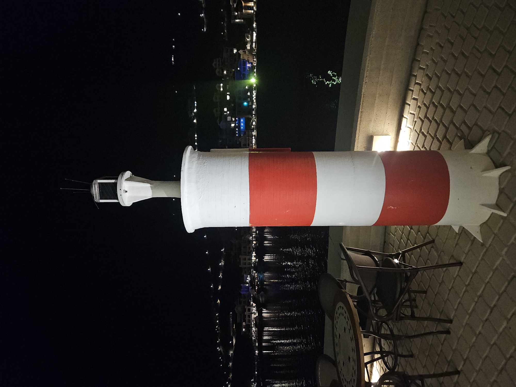 This is a nighttime outdoor photograph taken at eye-level. In the foreground, a red and white striped lighthouse stands upright on a paved surface. The lighthouse has a conical top with a lantern room. To the left of the lighthouse, a circular table with a patterned mosaic top is set with two chairs. The chairs are made of dark metal with woven seats and backs. In the background, a body of water reflects the lights of a town or village that stretches along a hillside. The lights are scattered and appear to be from buildings and streetlights. The sky is dark and clear. The scene suggests an outdoor seating area overlooking a harbor or bay at night.