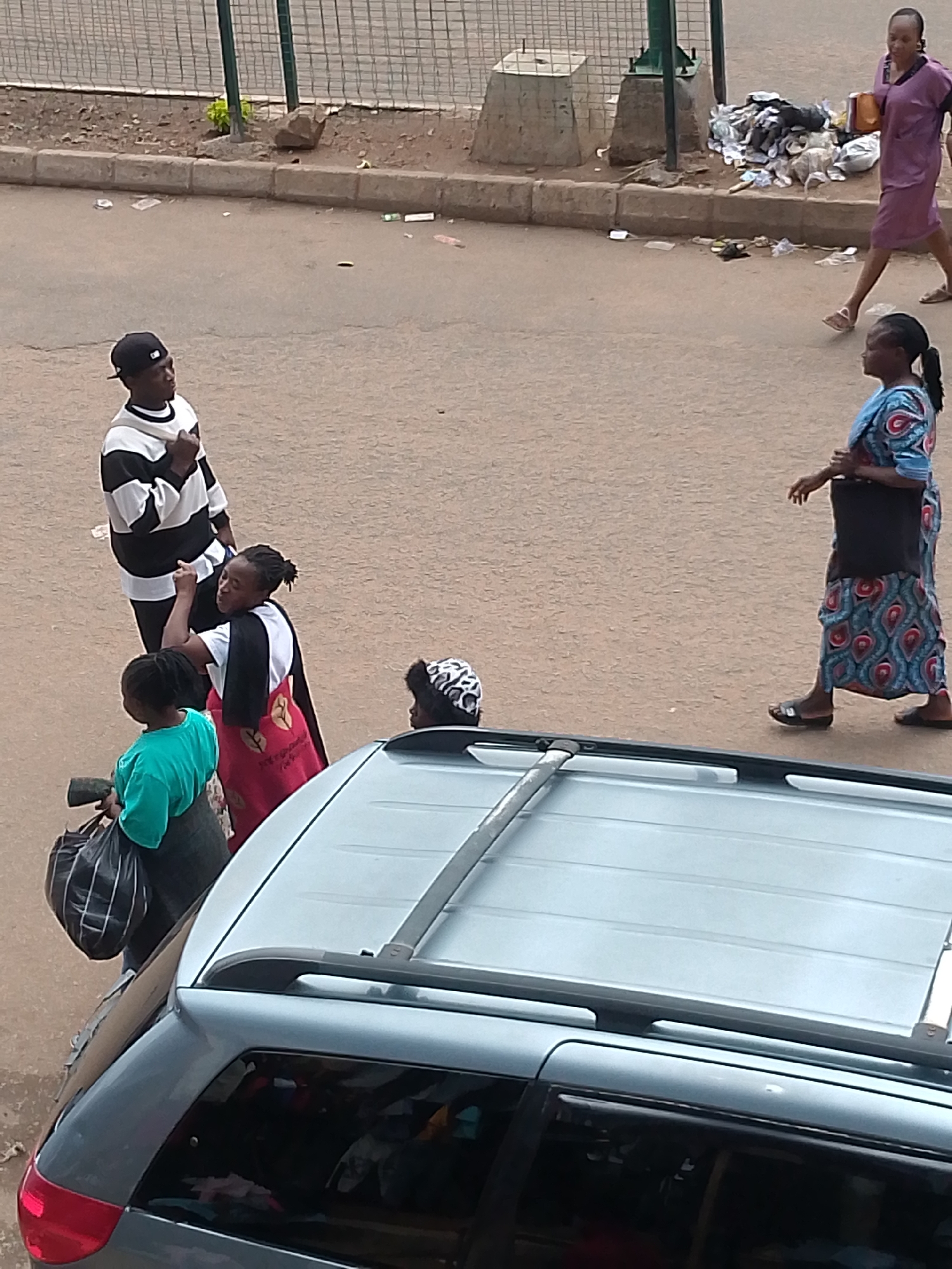 The image shows a street scene in Bukuru, Nigeria, with several people and a vehicle. In the foreground, a silver car is parked. In the middle ground, a man in a black and white striped shirt and black baseball cap is standing, facing left and gesturing with his right hand. To his left, a woman wearing a green shirt is holding a black bag. Behind them, a woman in a red and black patterned skirt and white top is facing the camera and appears to be speaking. Further to the right, a woman in a blue and red patterned dress with a black bag over her shoulder is walking away from the camera. In the background, a woman in a purple dress is walking past a pile of garbage. The ground is paved, and there is a fence and some vegetation in the background.