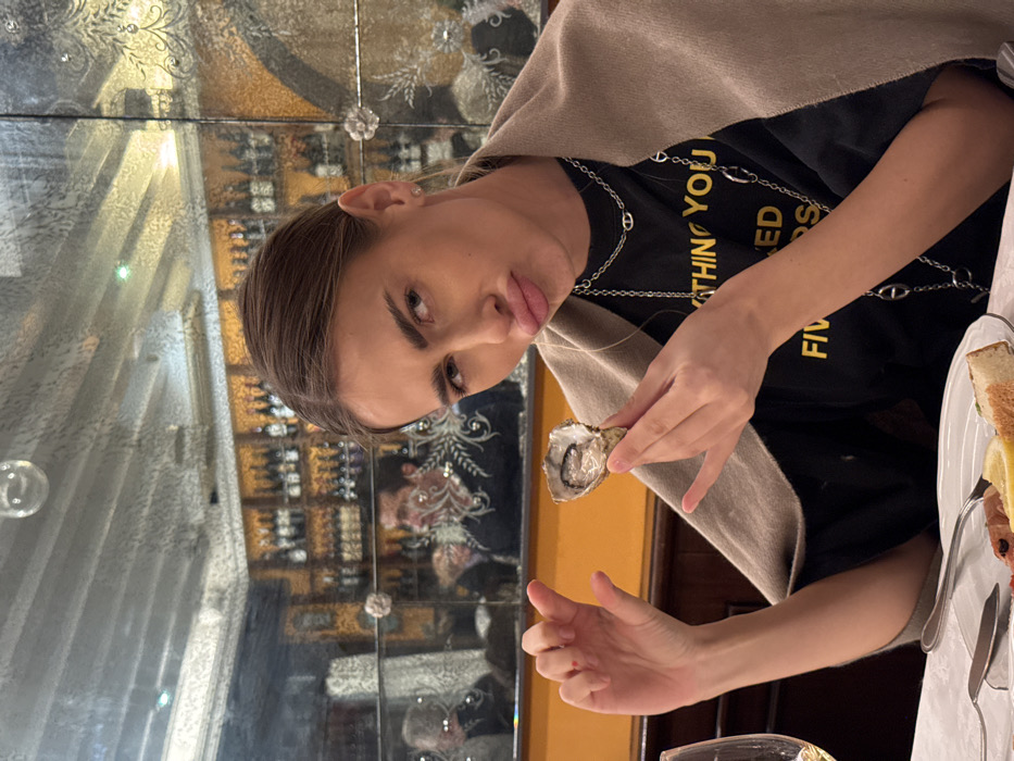 A young adult female is seated in the foreground of a traditional restaurant in Venice, Italy. Her light brown hair is pulled back, and she wears a black t-shirt featuring partially visible yellow text, multiple silver-toned chain necklaces, and a beige shawl draped over her shoulders. She holds an opened oyster in her right hand, displaying the bivalve meat, while her left hand is raised with fingers slightly splayed, showing a small red mark on the index finger. Her expression consists of pouted lips and raised eyebrows, directed towards the camera. On the white-tableclothed table in front of her are a white wine glass and a plate containing cutlery, a lemon wedge, a piece of bread, and a portion of cured meat. The background features ornate, segmented mirrored walls with etched floral patterns. These mirrors reflect the restaurant's interior, including shelves densely stocked with wine bottles, warm-toned walls, and other patrons, notably a male figure with dark hair and an embroidered sweater visible in the upper-left reflection. A small, clear spherical object is also reflected in the upper center of the mirrored surface.