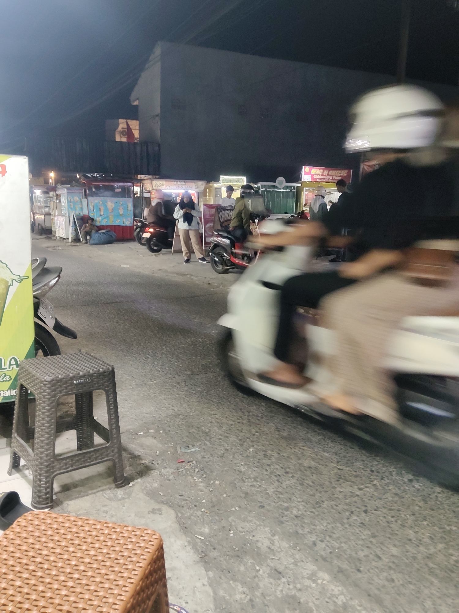 The image depicts a street scene at night in Mekarmukti, Indonesia, likely an area with food vendors. In the foreground, two plastic stools are visible on a paved road. A dark brown stool with a woven texture is on the left, and a lighter brown, taller stool with a more intricate woven pattern is closer to the center. The background shows several food stalls with various colorful signs, illuminated by artificial lights. People are seen at and around the stalls, some ordering food, others standing or walking. Several motorbikes are parked or in motion. Notably, a person wearing a white helmet is riding a motorbike, moving from right to left across the frame, their image blurred due to motion. The overall atmosphere is that of a bustling evening marketplace.