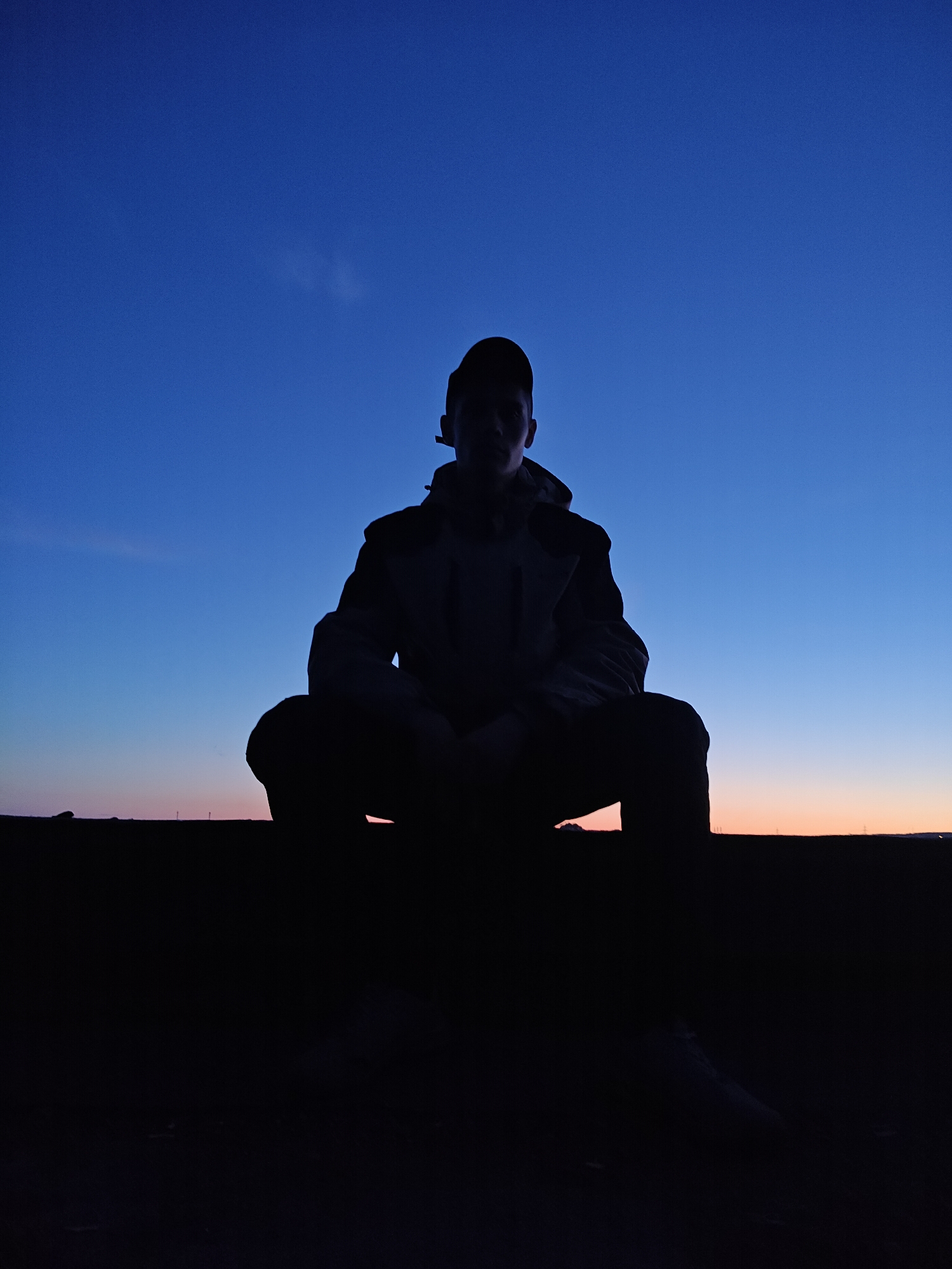The image features a solitary figure, likely a person, captured in silhouette against a vibrant twilight sky. The scene is set outdoors, possibly in Chaikovsky, Russia, during either dusk or dawn.
The central subject, a person, is shown in a low-crouch or squatting position. They are wearing a cap, pulled low over their face, and a jacket, which has some subtle textural details visible despite the deep shadows. Their form is almost entirely silhouetted, appearing as a dark outline against the brighter background. Minimal light gently catches parts of their face, such as the forehead and cheek, hinting at their features but keeping them largely obscured.
The sky forms the dominant backdrop, exhibiting a striking color gradient. It transitions from a deep, rich blue at the top of the frame to lighter shades of blue, eventually blending into soft bands of orange, pink, and pale yellow along the horizon line. This clear, cloudless sky indicates a serene atmospheric condition.
The lower portion of the image, below the horizon, is rendered in complete darkness, suggesting a foreground object or ground that is heavily shadowed. The distinct horizontal line separating the dark foreground from the illuminated sky implies an open landscape or a distant elevated edge. The overall mood conveyed is one of quiet contemplation or a posed moment against a beautiful natural light display. No visible text is present in the media file.