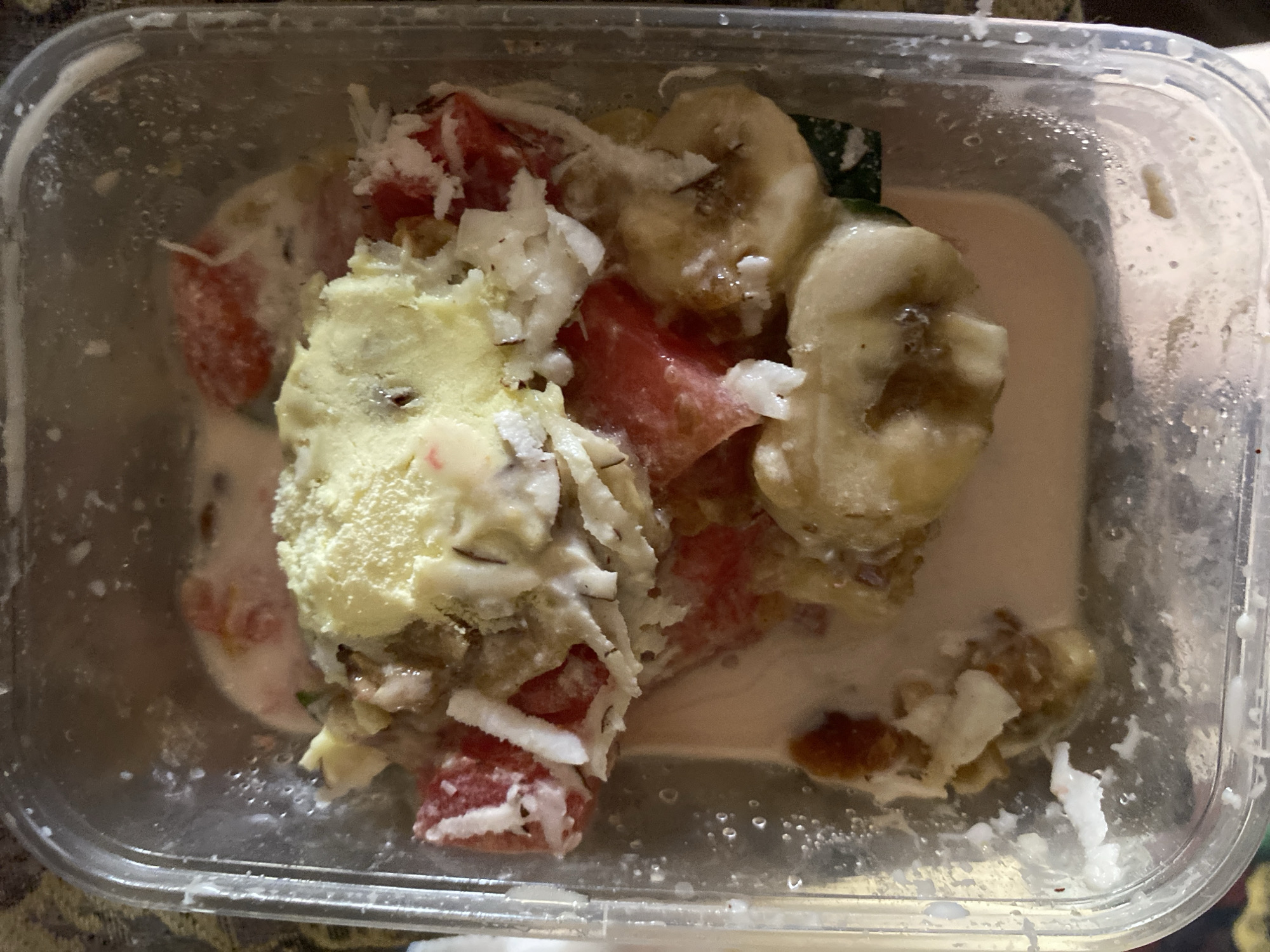 A clear plastic container holds a mixture of fruit and creamy liquid. The fruits visible include sliced bananas, chunks of watermelon, and a scoop of what appears to be ice cream or custard. Shredded coconut is sprinkled throughout the mixture. The container is partially filled with a light-colored, milky liquid. The container's exterior shows condensation. The scene is indoors. The location, Barakin Akawo, Nigeria, is not visually identifiable in the image.