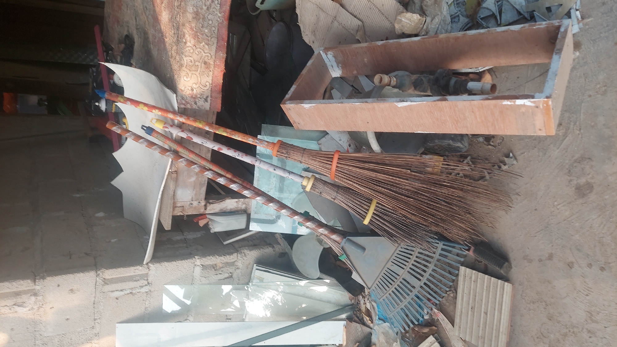 This is a forensic, matter-of-fact description of a scene in Restu Baru, Indonesia.
The image depicts a cluttered outdoor area, likely a storage or work space, with a concrete floor. In the foreground, three brooms are propped up, leaning against each other and a rake. The brooms have natural fiber bristles and decorated wooden handles with visible patterns and colors. One broom has a yellow band around its bristles, and another has an orange band. A blue and grey rake with a metal head and plastic handle is positioned to the left of the brooms.
Behind the cleaning tools, a large, white, curved piece of material, possibly plastic or cardboard, is supported by a makeshift wooden structure. To the left of this structure, several panes of glass are leaning against a rough, concrete block wall. Some of these glass panes are transparent, while others appear to have reflective surfaces.
Further in the background, various discarded materials are visible, including corrugated metal sheets, cardboard, and possibly buckets. A partially visible wooden frame with some internal components, possibly plumbing or electrical fixtures, is present in the right foreground. The overall environment appears to be disorganized and under a bright, natural light source, suggesting daytime. There are no people visible in the image.