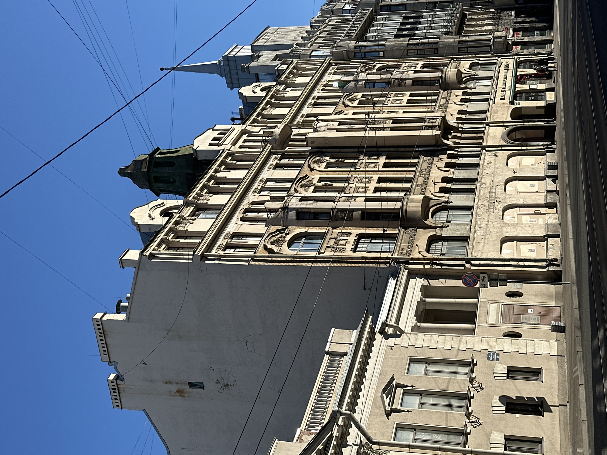 The image depicts a tall, ornate building in an urban environment. The building has multiple floors with a variety of architectural details, including windows, balconies, and decorative elements. There are visible power lines in the foreground. The sky is clear and blue. The perspective is from below, looking upward at the building. A street is partially visible on the right. The location is in the city of Unknown, Unknown.
