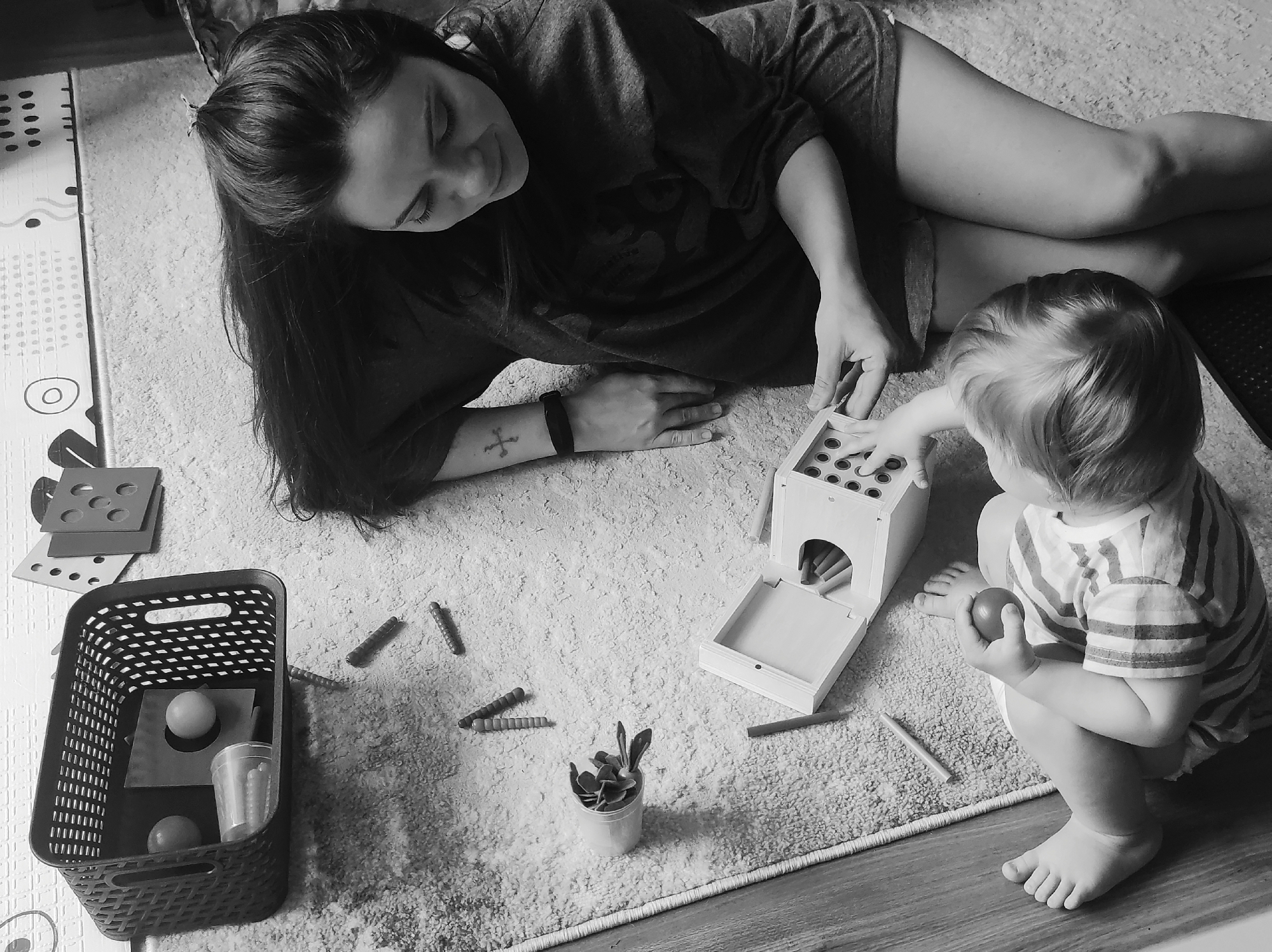 An overhead, black and white photograph captures an intimate indoor scene in what appears to be a home in Chaikovsky, Russia. An adult woman and a young child are engaged in playtime on a light-colored, shaggy rug.
The woman lies on her stomach on the left side of the frame, looking down with a gentle expression towards the child and the toys. She has long, dark hair and wears a dark t-shirt and shorts. A cross tattoo is visible on her left forearm.
The child, with blondish hair and wearing a striped t-shirt, sits opposite her on the right. The child is holding a red ball and actively interacts with a wooden educational toy, possibly a shape sorter or peg box, which has multiple holes and a pull-out drawer. Several crayons or pegs are scattered around the toy and on the rug, with some also contained in a small white cup.
Other play items are visible, including a black plastic woven basket containing another ball, a black square, and a clear cup, along with several square-shaped puzzle pieces or cards in the upper left corner. A patterned play mat is partially visible beneath the rug on the left, and a wooden floor borders the rug on the right. The overall atmosphere suggests a quiet, focused, and tender moment of shared activity between the adult and child. No discernible text is visible on clothing or objects.